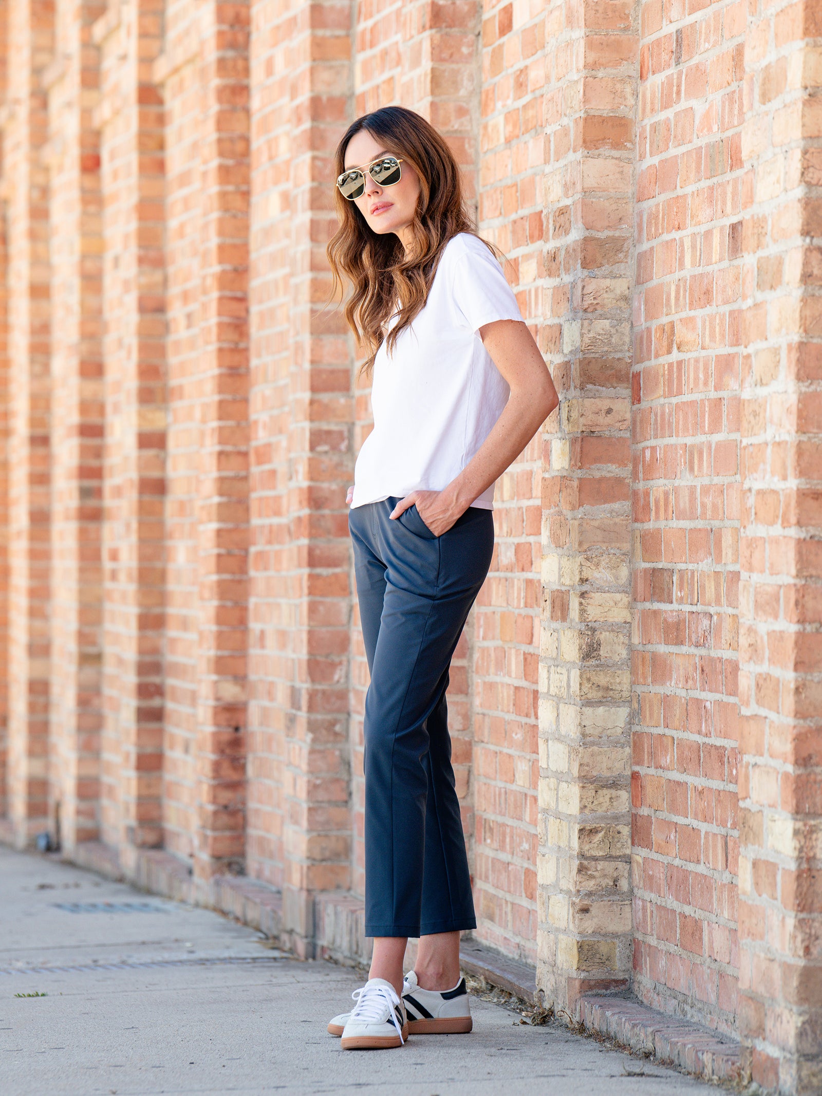 A person with long hair wearing sunglasses, a white t-shirt, Cozy Earth's Women's Always Cropped Pant, and sneakers is standing against a brick wall. They have their hands in their pockets and are gazing into the distance. 