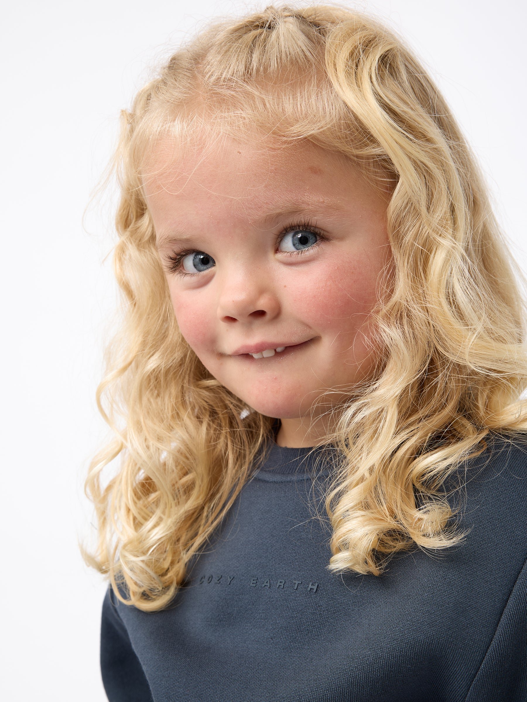 A young child with curly blonde hair smiles slightly, wearing the Kid's CityScape Crewneck from Cozy Earth, featuring a dark gray design with subtle text. The background is plain white. 