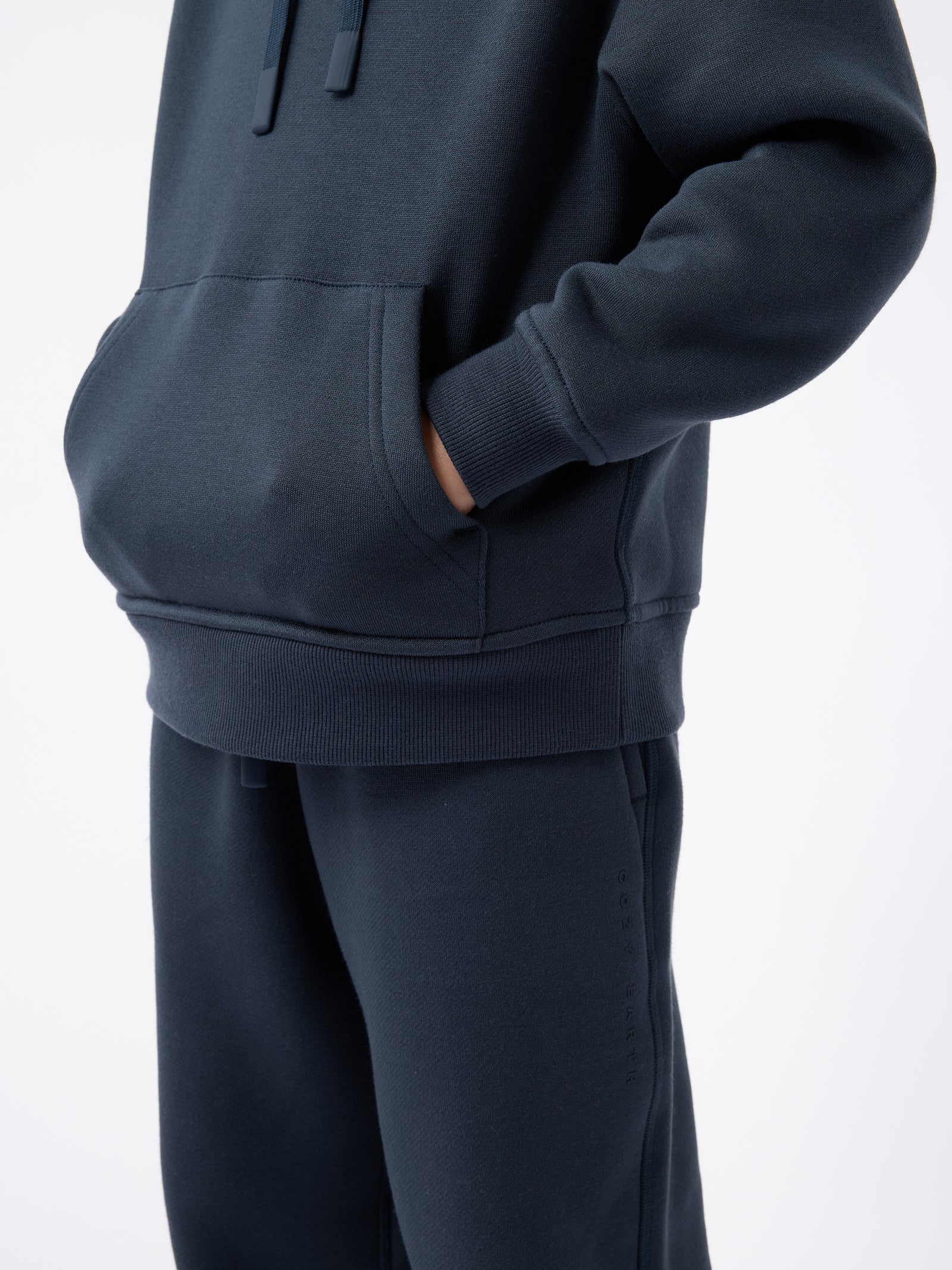 A person wearing the Kid's CityScape Hoodie by Cozy Earth, featuring a minimalist design in navy blue. The outfit includes the hoodie and matching pants, with their hands tucked into the front pocket. The background is plain white. 