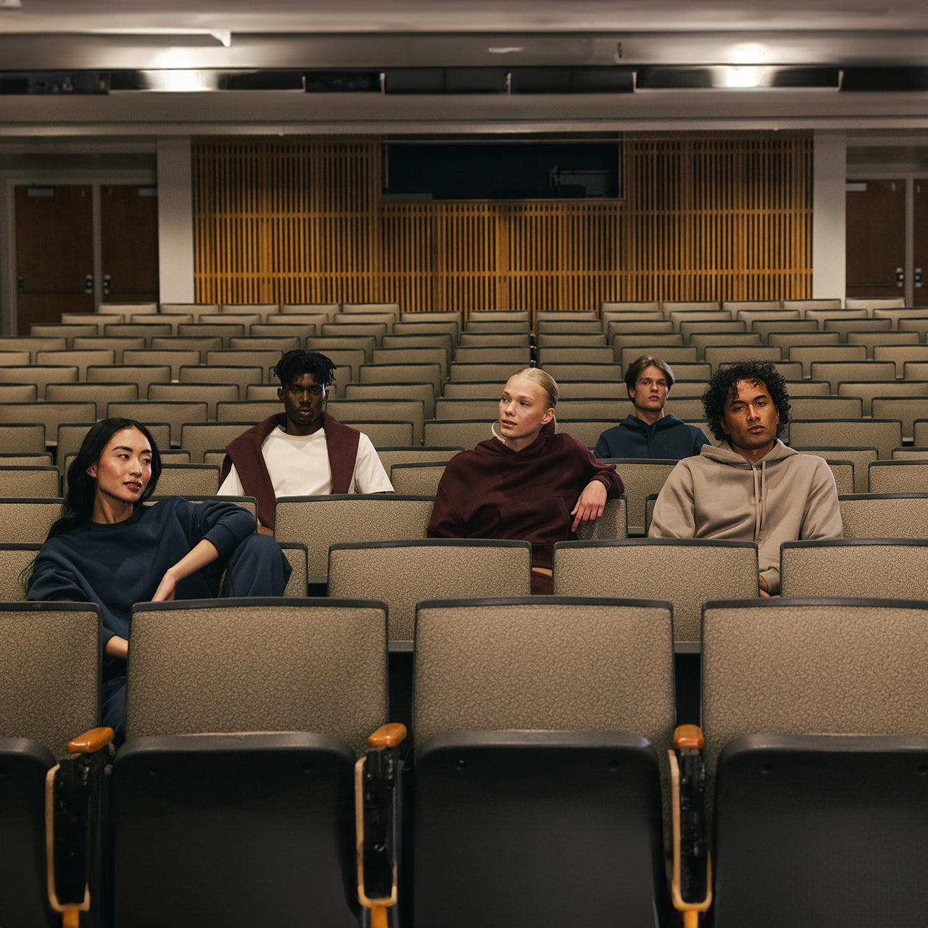 Five people are sitting spread out in an empty lecture hall with gray seats and light wood paneling on the back wall. They are casually dressed, some of them wearing the Women's CityScape Crewneck from Cozy Earth, each looking in different directions and giving the scene a contemplative atmosphere. 