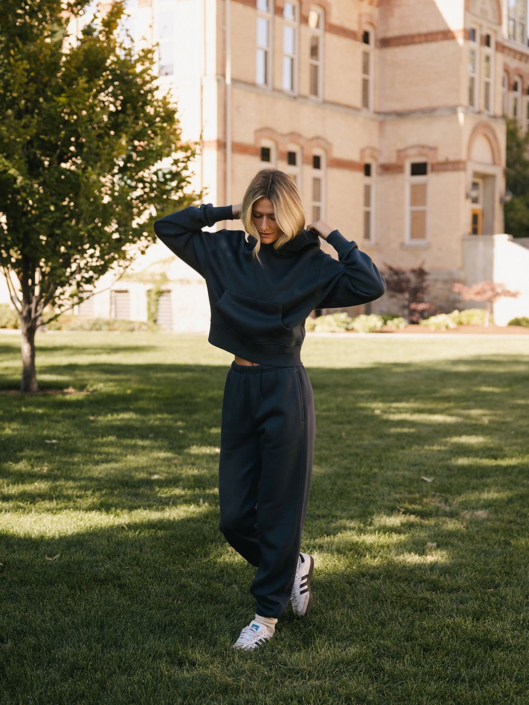 A person with shoulder-length blonde hair is standing on grass in front of a large brick building with tall windows. They are wearing a dark CityScape Cropped Hoodie by Cozy Earth, matching pants, and white sneakers, with their hands raised to adjust their hair or clothing. 