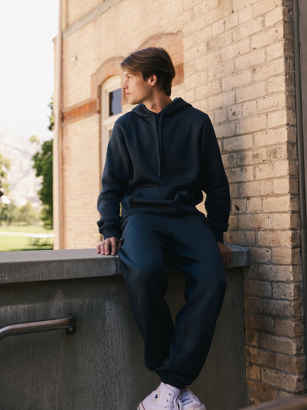 A person wearing a Cozy Earth Men's CityScape Hoodie and sweatpants sits on the edge of a concrete railing, looking to the side. The background features a brick building and trees, with a glimpse of a mountainous landscape. The person is also wearing white sneakers. 