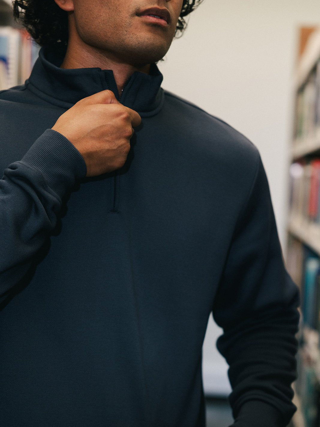 A person with curly hair is depicted from the nose down, wearing a dark blue Men's CityScape Quarter Zip sweater by Cozy Earth. They are adjusting the zipper with one hand. The background features blurred shelves filled with books, suggesting a library or bookstore setting. 