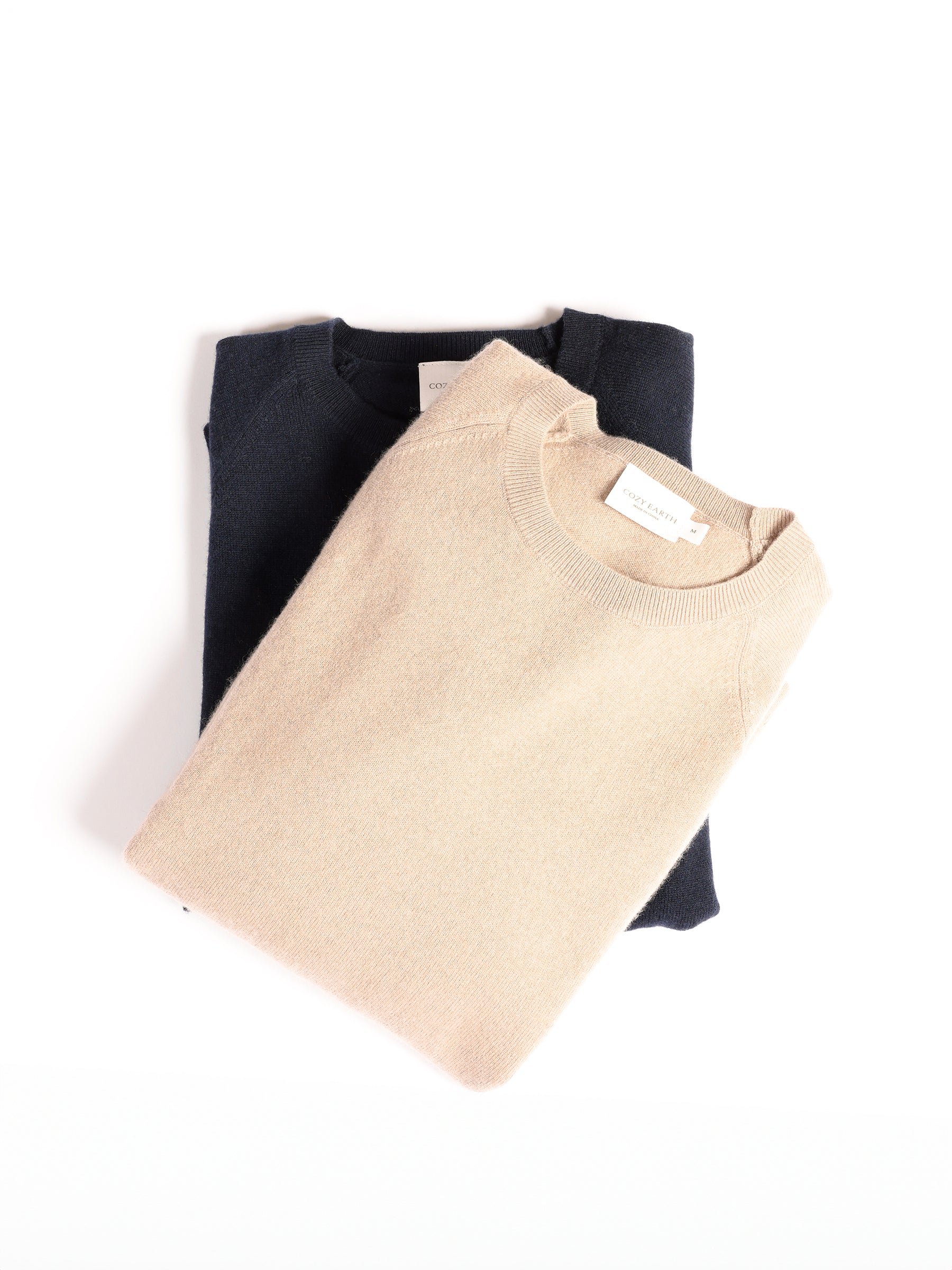 Two neatly folded Men's Crewneck Sweaters by Cozy Earth are placed on a white background. One sweater is beige with a visible label at the neckline, and the other one underneath is dark blue. The sweaters are arranged slightly overlapping. 
