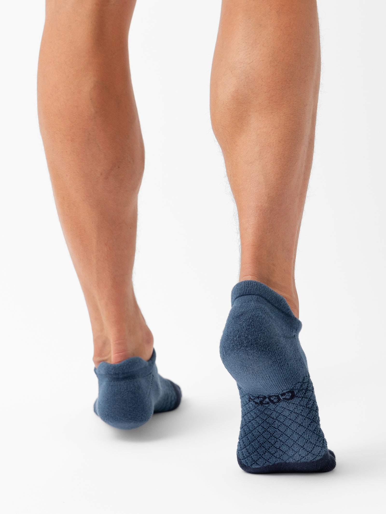 Rewritten Sentence: Rear view of a person wearing Cozy Earth's Essential Ankle Socks in blue, displaying their lower legs and feet. The socks feature a textured pattern on the soles and a ribbed design around the ankles. The background is white. 