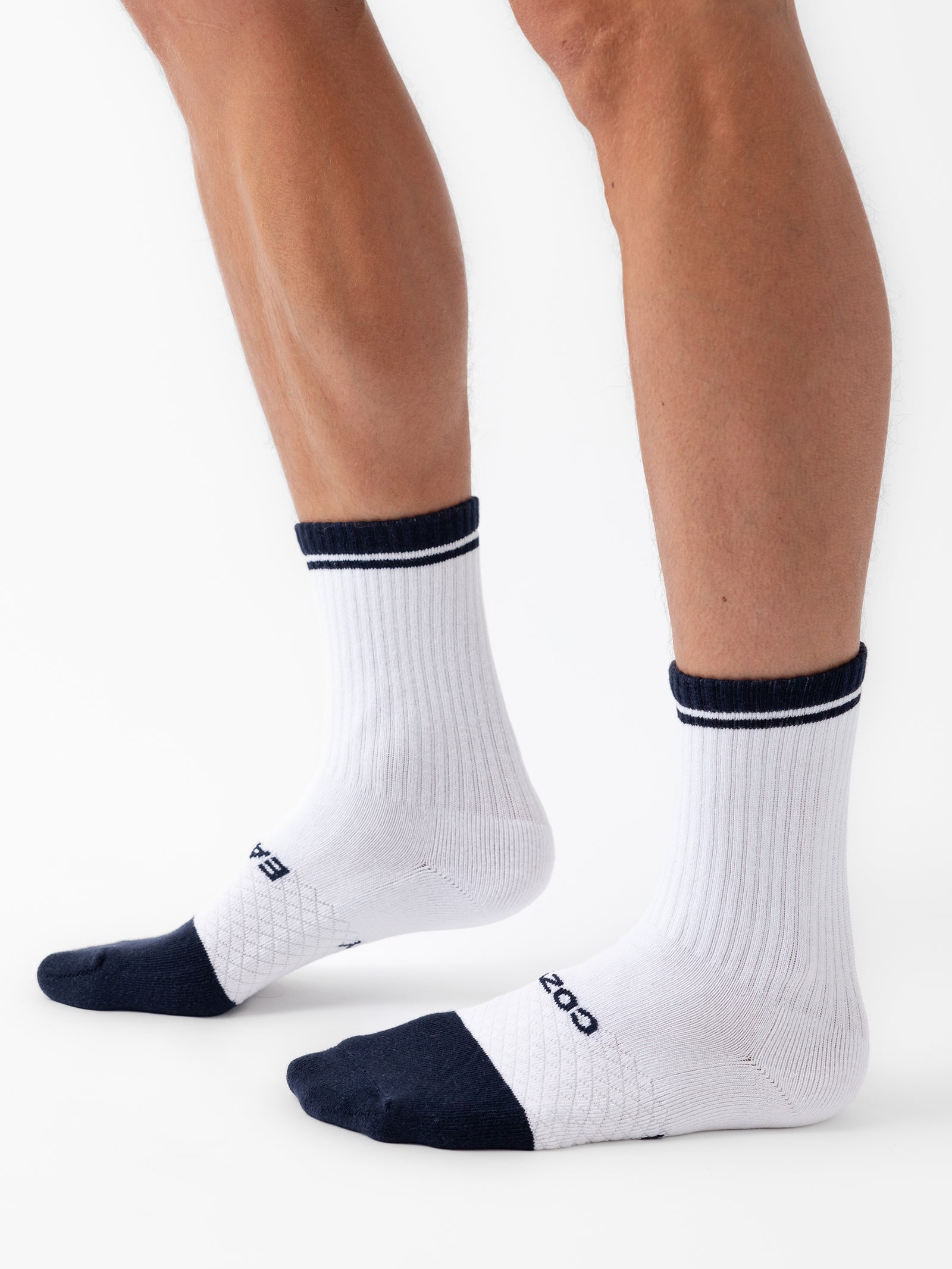 Person wearing white Essential Calf Socks from the Cozy Earth 2-Pack collection, standing on a white background. The socks feature a ribbed texture with the word "BOMBAS" written near the toes and have black toes and cuffs. 