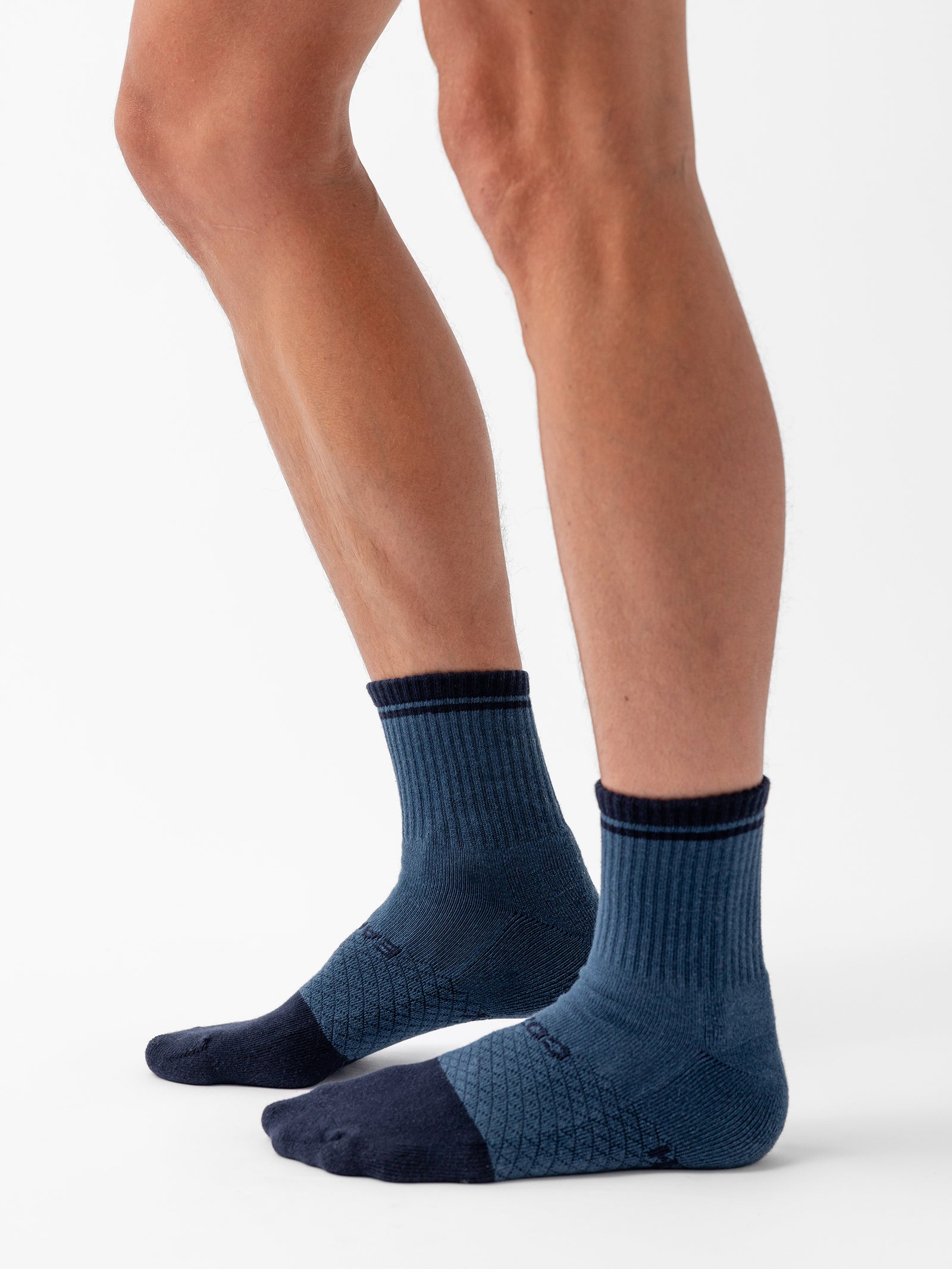 The image features a person's legs adorned with a pair of dark blue Essential Quarter Socks from Cozy Earth. These socks exhibit a ribbed upper section and a textured pattern on the foot area, complemented by darker toe and heel sections. The background is plain white. 