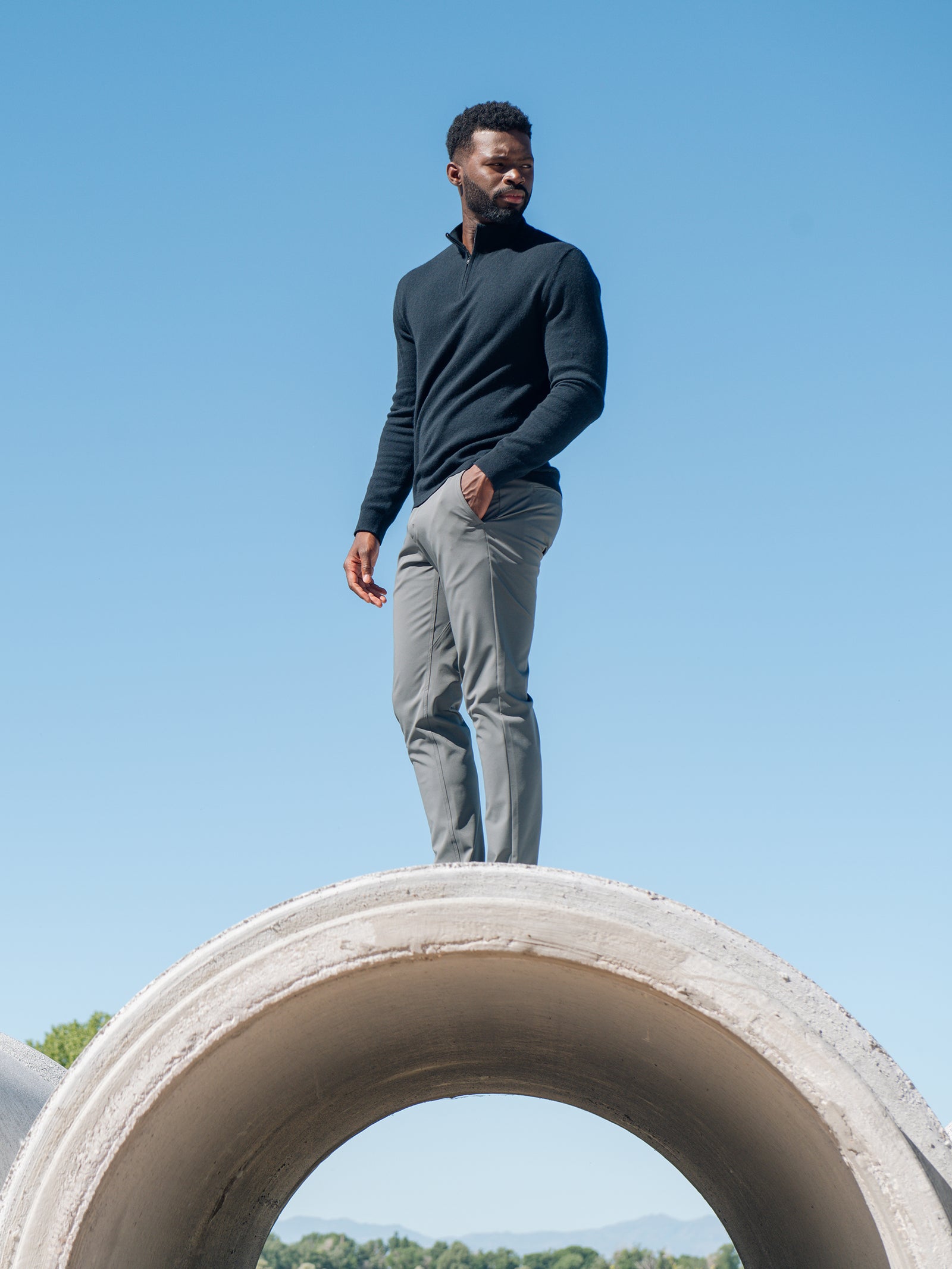 A man stands confidently on a large concrete pipe against a clear blue sky. He is wearing the Cozy Earth Men's Quarter Zip Sweater and gray pants. Trees and mountains are visible in the distant background, highlighting the open, outdoor setting. 