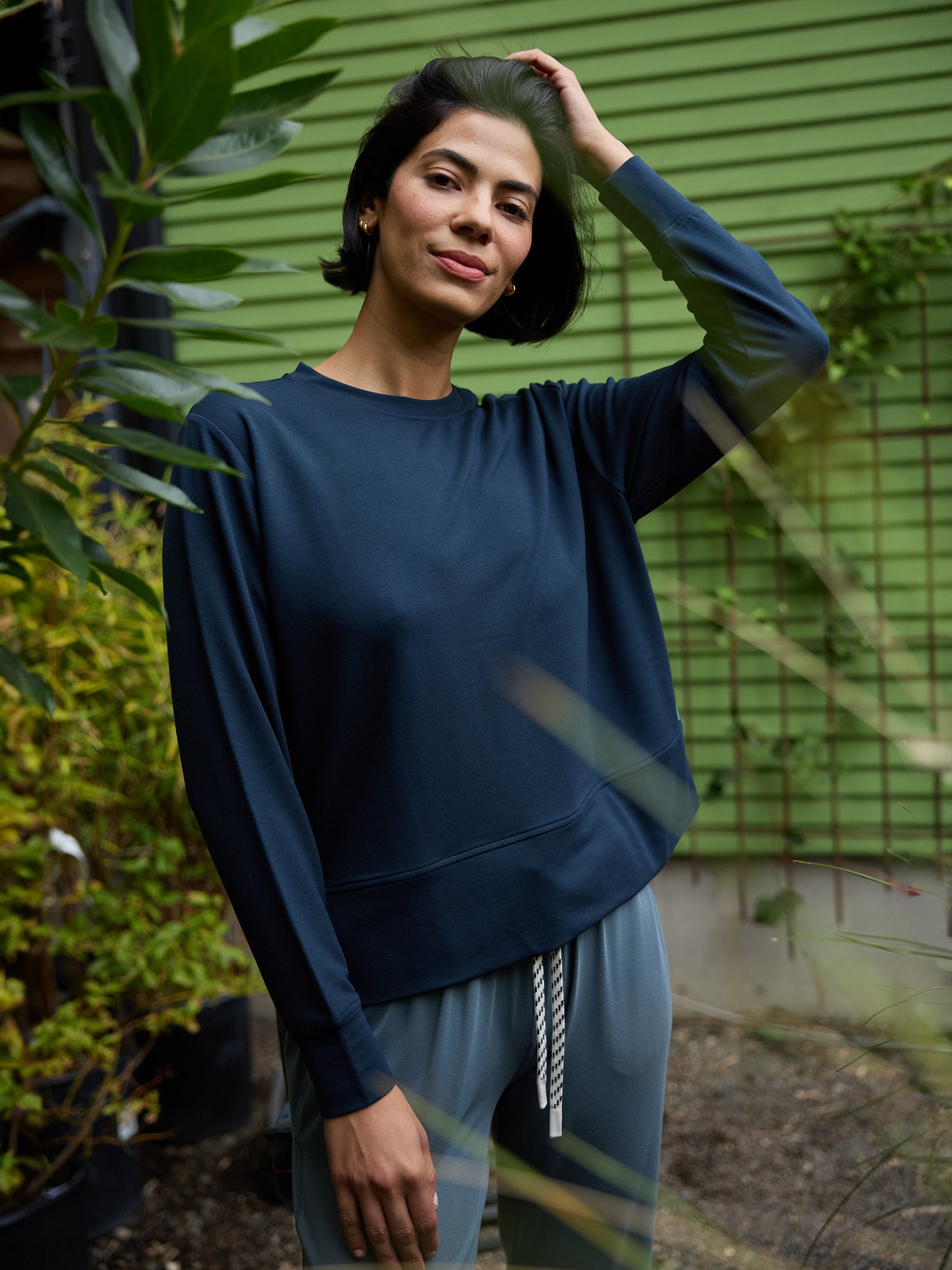 A person with short black hair stands outdoors in front of a green wall, wearing a navy blue Women's StudioLite Crewneck by Cozy Earth and light blue pants. They have one hand on their head and are smiling slightly. The background includes plants and garden elements. 