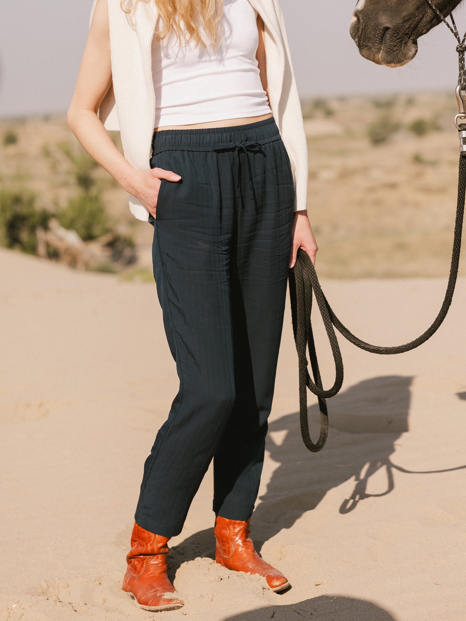 A person with long, wavy hair is standing on a sandy surface holding a horse's reins. They are wearing a sleeveless white top, Cozy Earth's Women's Sunset Cropped Pant, and orange boots. The scene is set in a desert-like area with sparse vegetation in the background. 