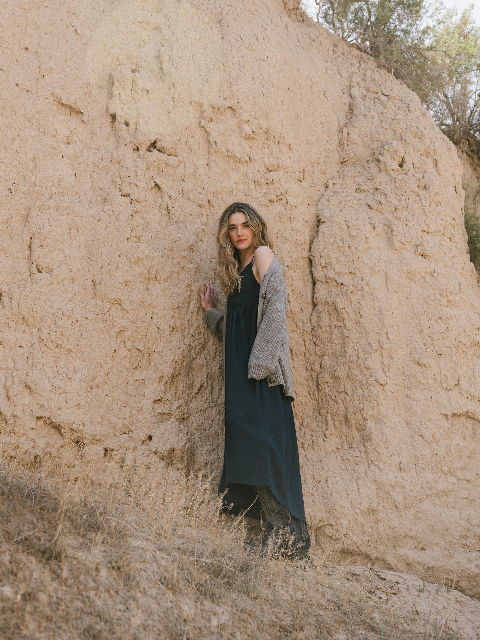 A person with long wavy hair, adorned in Cozy Earth's Women's Sunset Gathered Dress and a gray cardigan, leans against a large, textured rock formation. The scene is outdoors with dry grass and a clear sky. The natural colors of the rock and landscape create a serene and earthy atmosphere. 