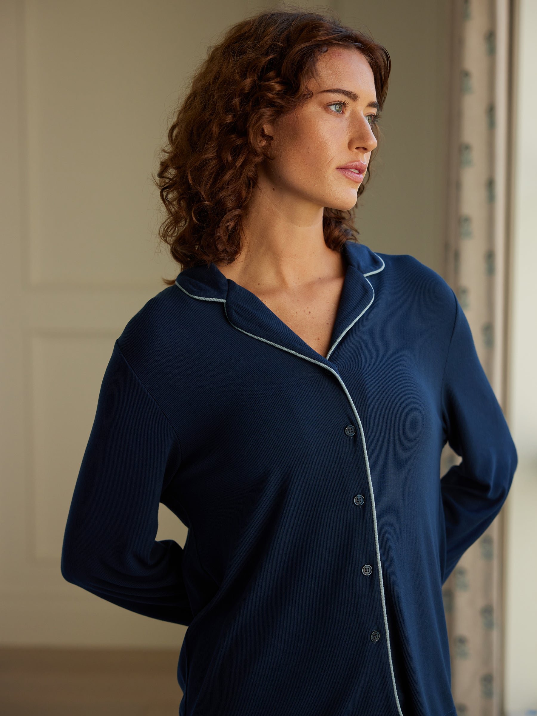 A person with curly hair, wearing a Cozy Earth Women's Bamboo Rib Knit Classic Long Sleeve Pajama Top in navy blue, stands indoors. They gaze out of a window to the right, with their hands resting behind their back. The background features light-colored walls and a partially visible curtain. 