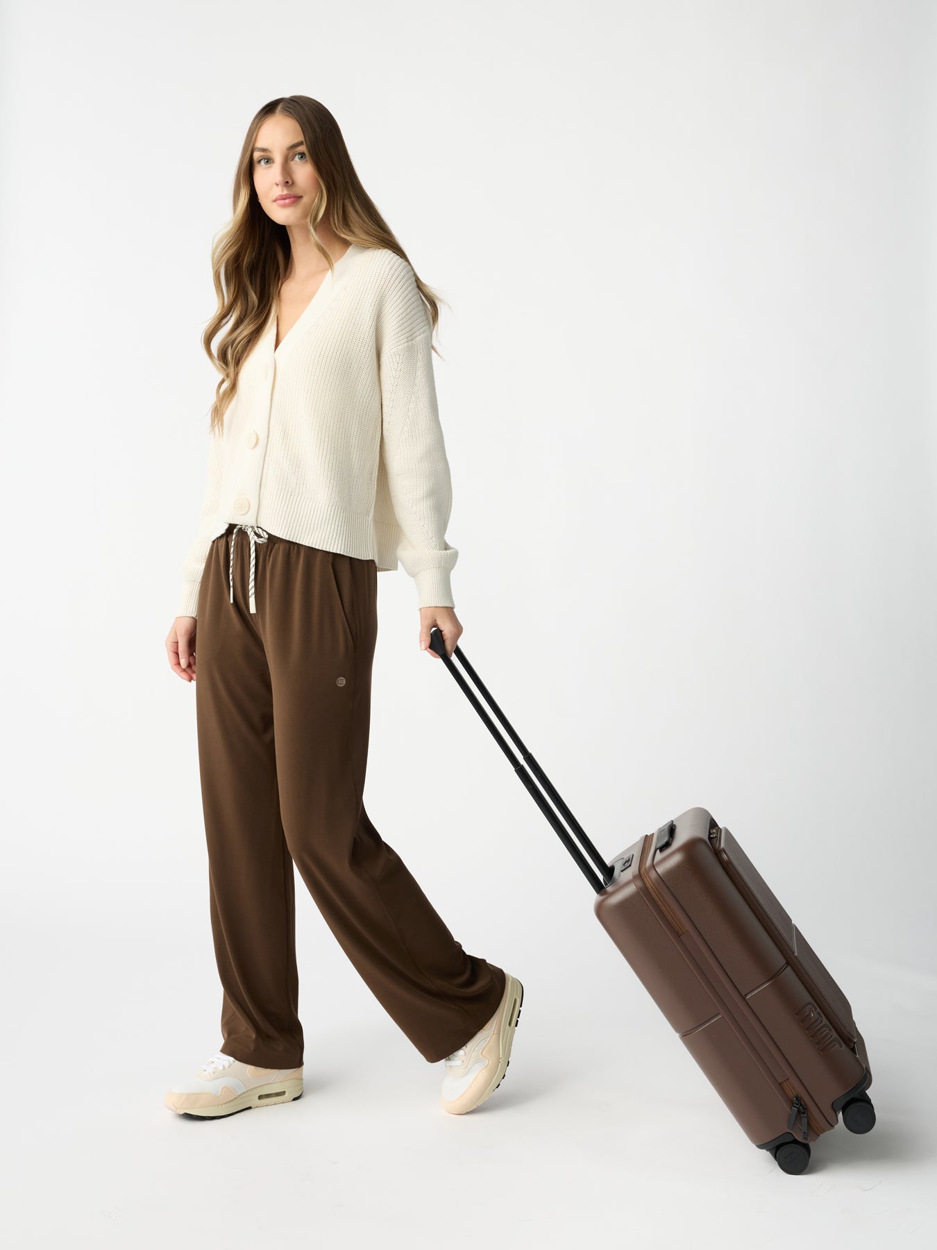 A woman with long hair stands against a white background wearing a cream-colored sweater, Women's Studio Wide Leg Pant by Cozy Earth, and cream sneakers. She is holding the extended handle of a brown suitcase with wheels. 