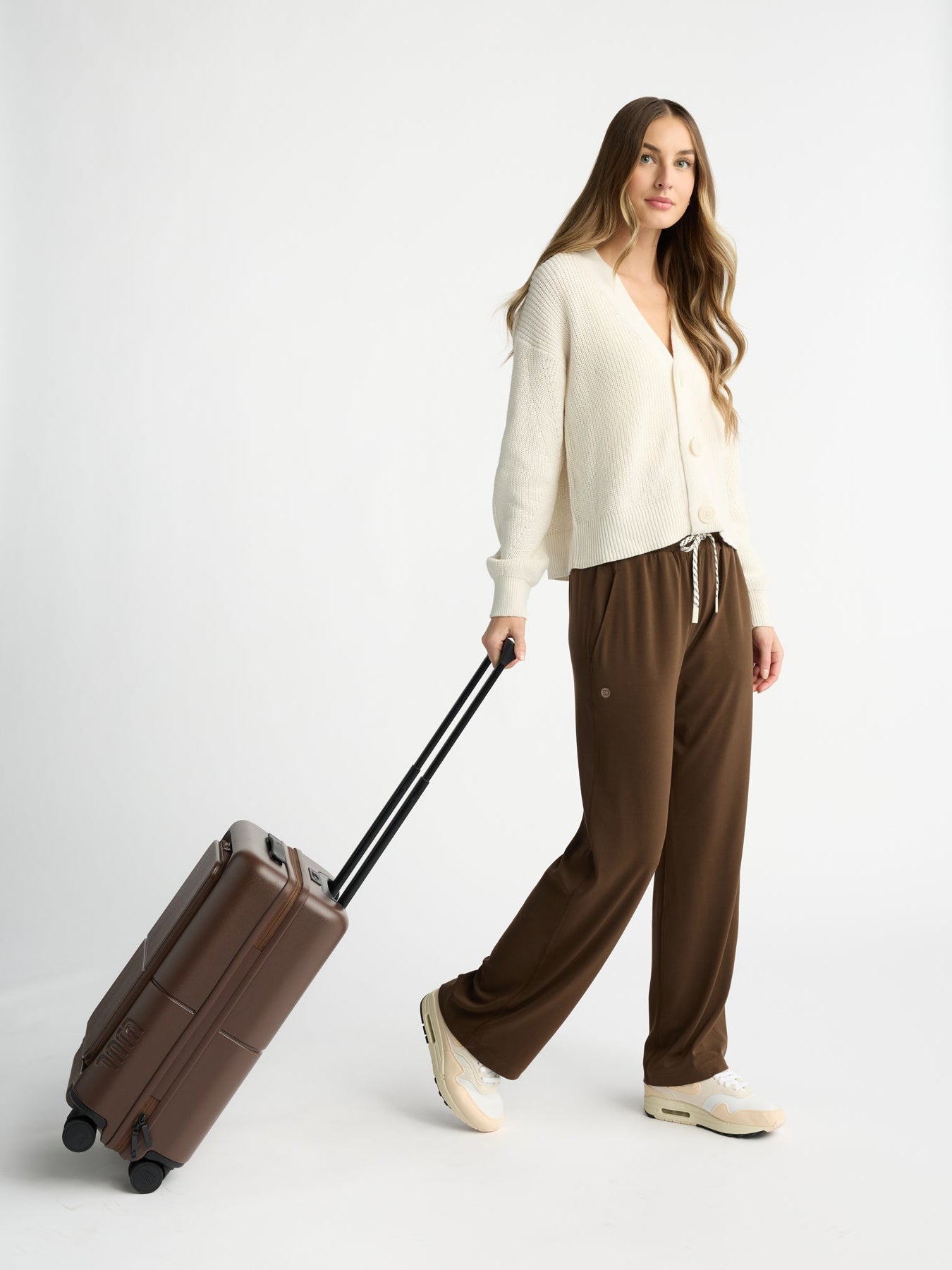 A woman with long hair stands against a white background wearing a cream-colored sweater, Women's Studio Wide Leg Pant by Cozy Earth, and cream sneakers. She is holding the extended handle of a brown suitcase with wheels. |Color:Espresso