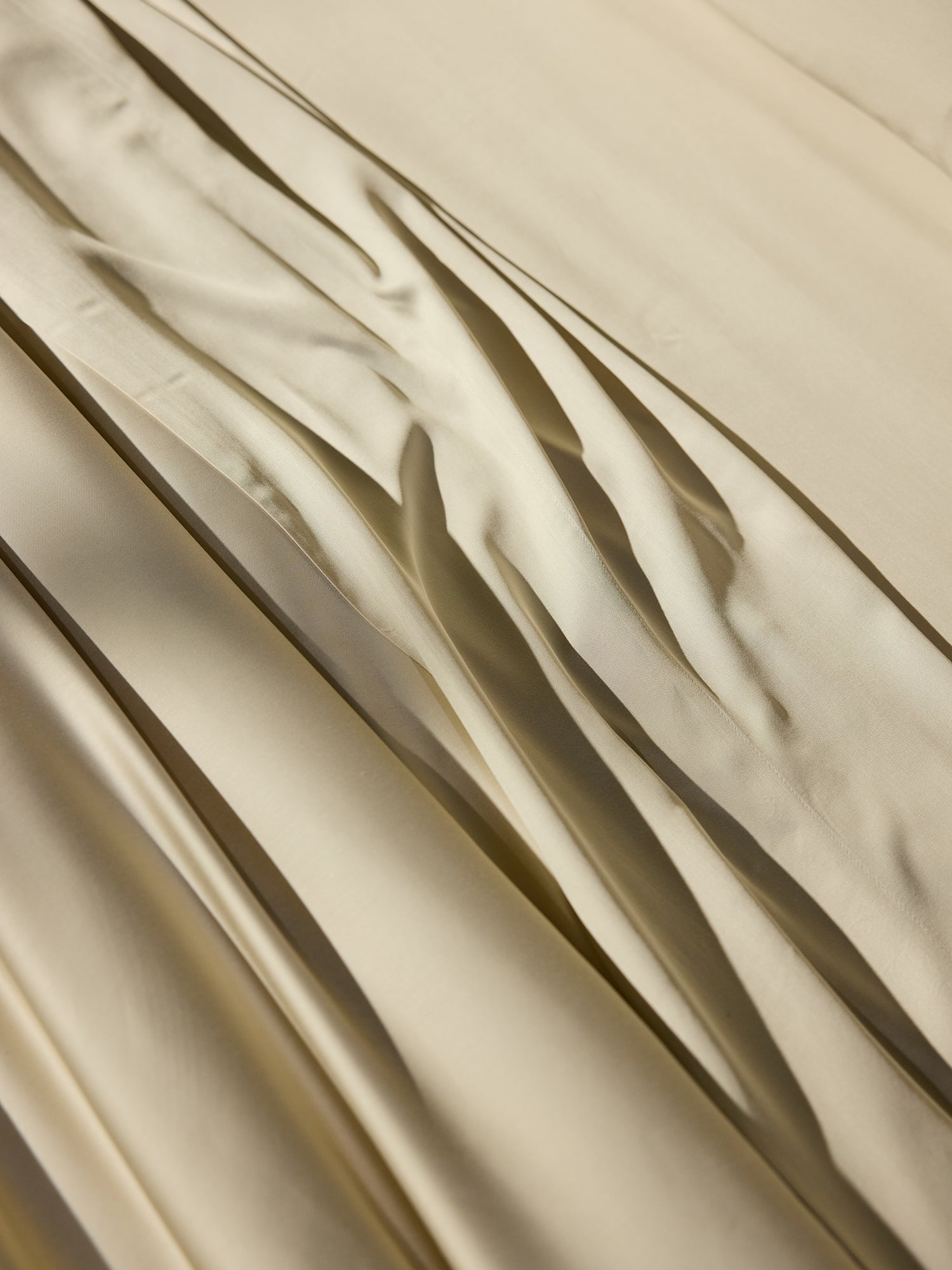 A close-up shot of Cozy Earth's Bamboo Fitted Sheet showcases its smooth, cream-colored fabric with gently flowing, overlapping folds, creating an undulating texture and a play of light and shadow on the surface. The sheet appears silky and soft. 