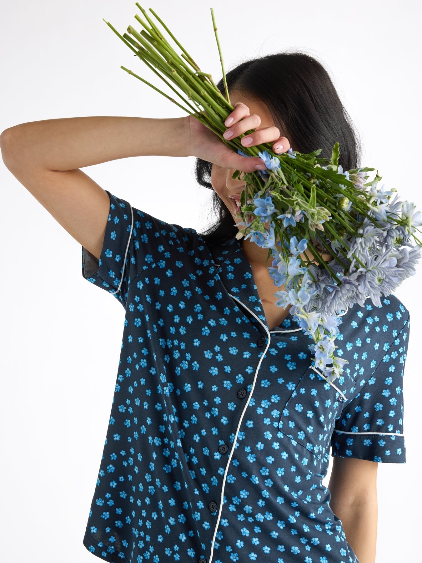 A person wearing HIDE's dark blue Women's Bamboo Stretch-Knit Short Sleeve Pajama Top with light blue floral patterns holds a bouquet of light purple flowers that partially covers their face against a plain white background. 