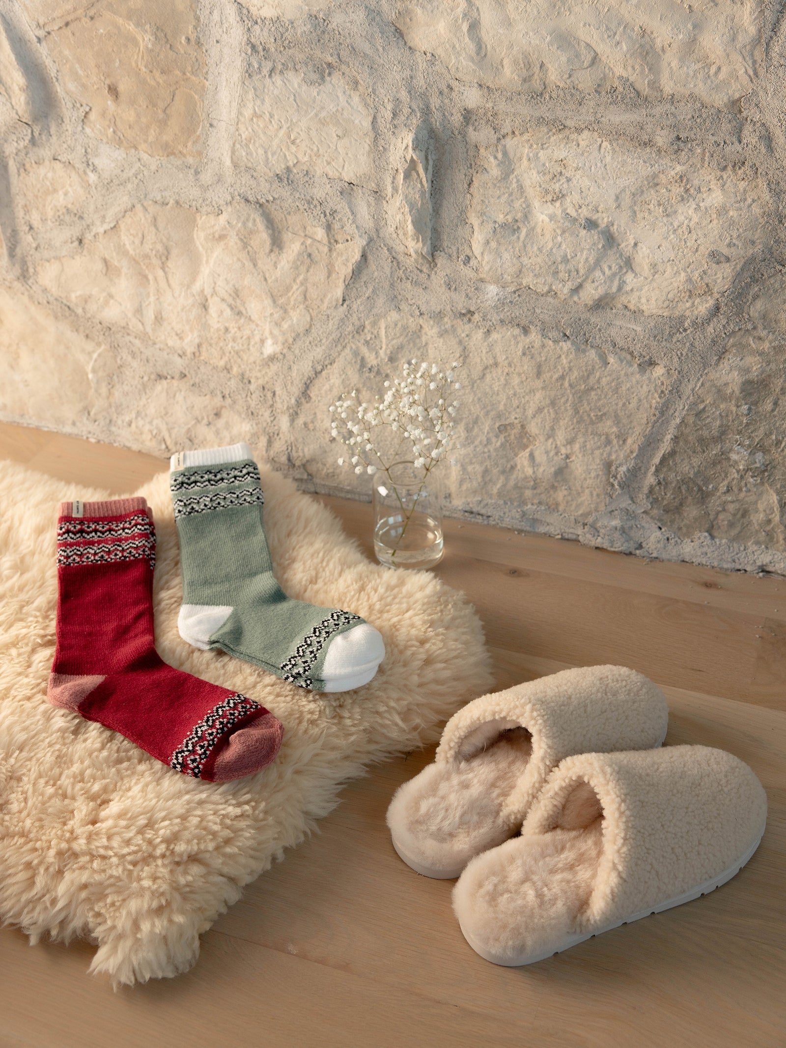 A pair of Fair Isle Plush Lounge Socks in Oakmoss and Garnet rest on a sheepskin rug near a pair of slippers and vase of flowers. 