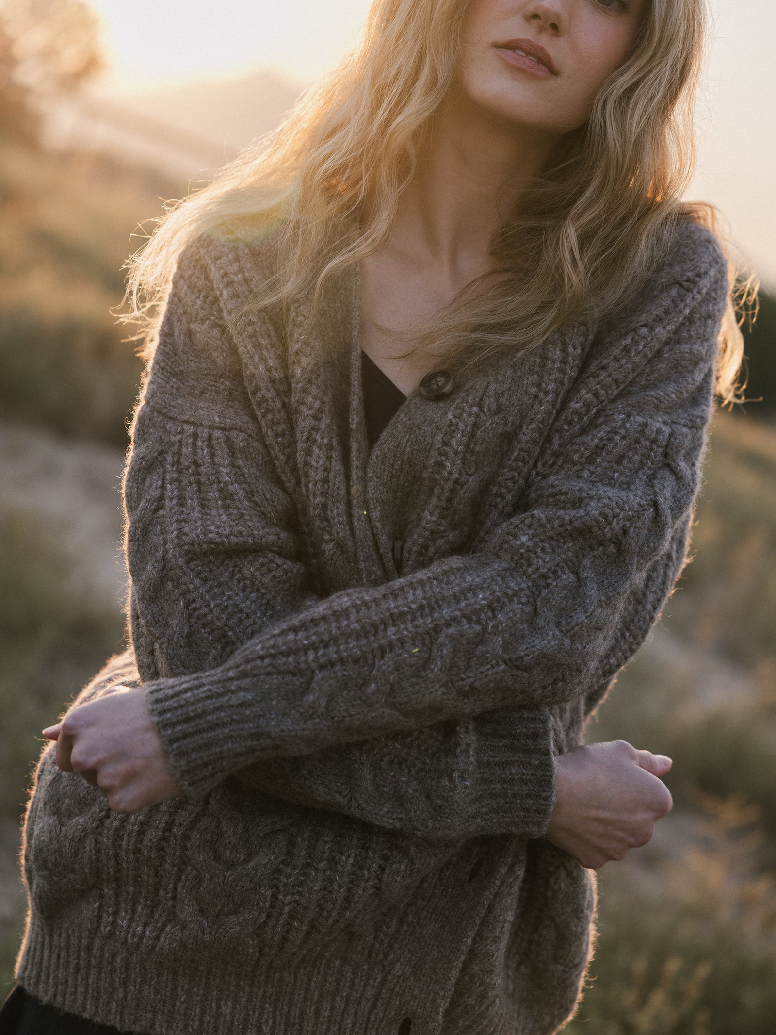 A person with long, wavy hair stands outdoors in warm lighting, wrapped in a Cozy Earth Oversized Cable Knit Cardigan. The background reveals a blurred natural setting, evoking a tranquil, autumnal ambiance. 