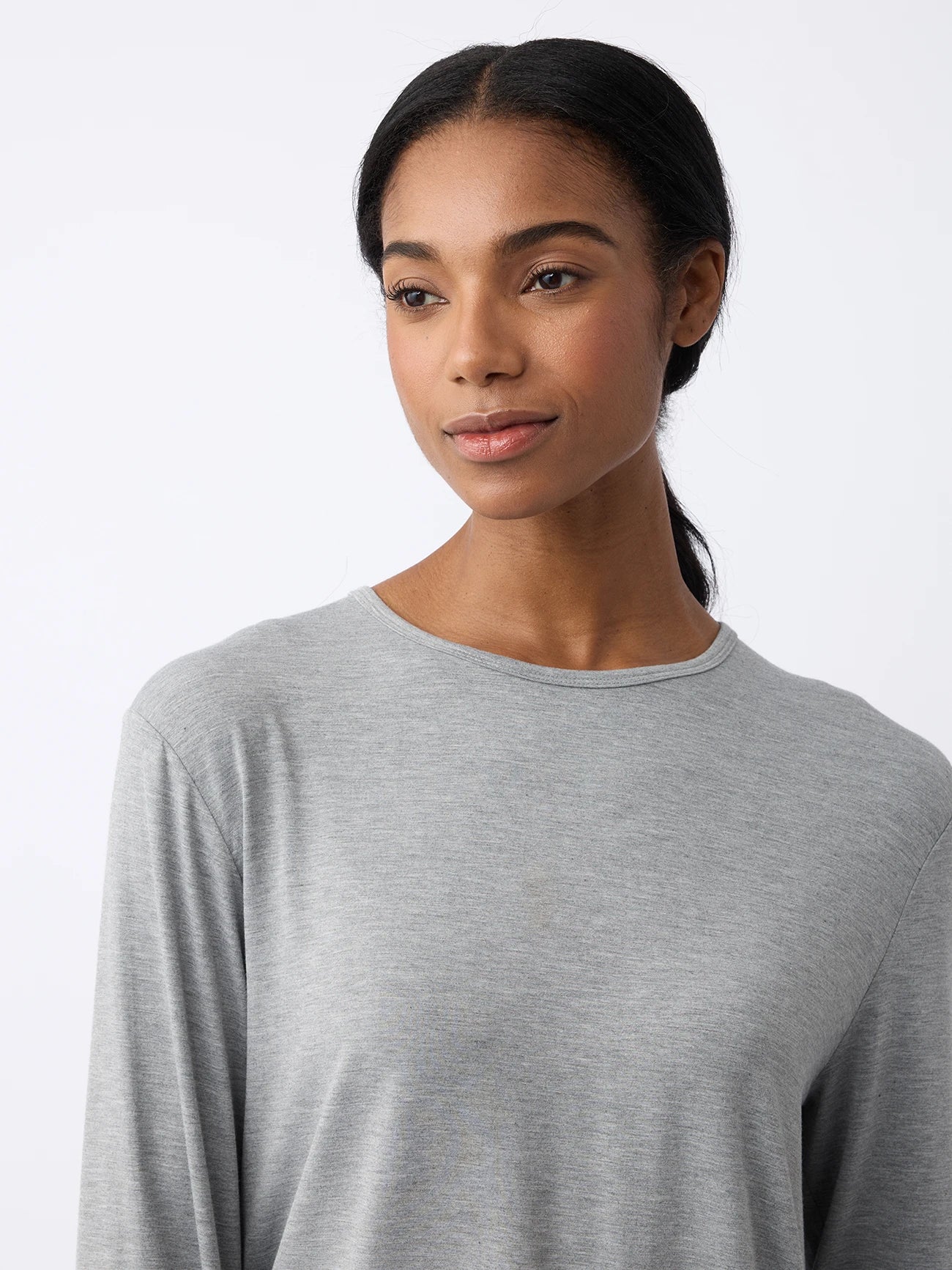 A long-haired person wearing a gray Women's Stretch Knit Bamboo Long Sleeve Lounge Tee from Cozy Earth looks to the side against a plain, light-colored background. 