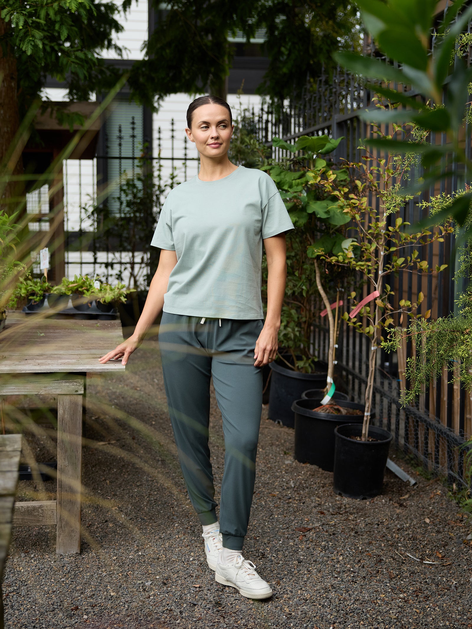 A woman stands outdoors near a wooden table and potted plants, wearing the Cozy Earth Women's All Day Tee in light green, along with dark green sweatpants and white sneakers. The background features trees and a fence adorned with additional greenery. She appears relaxed and is smiling gently. 