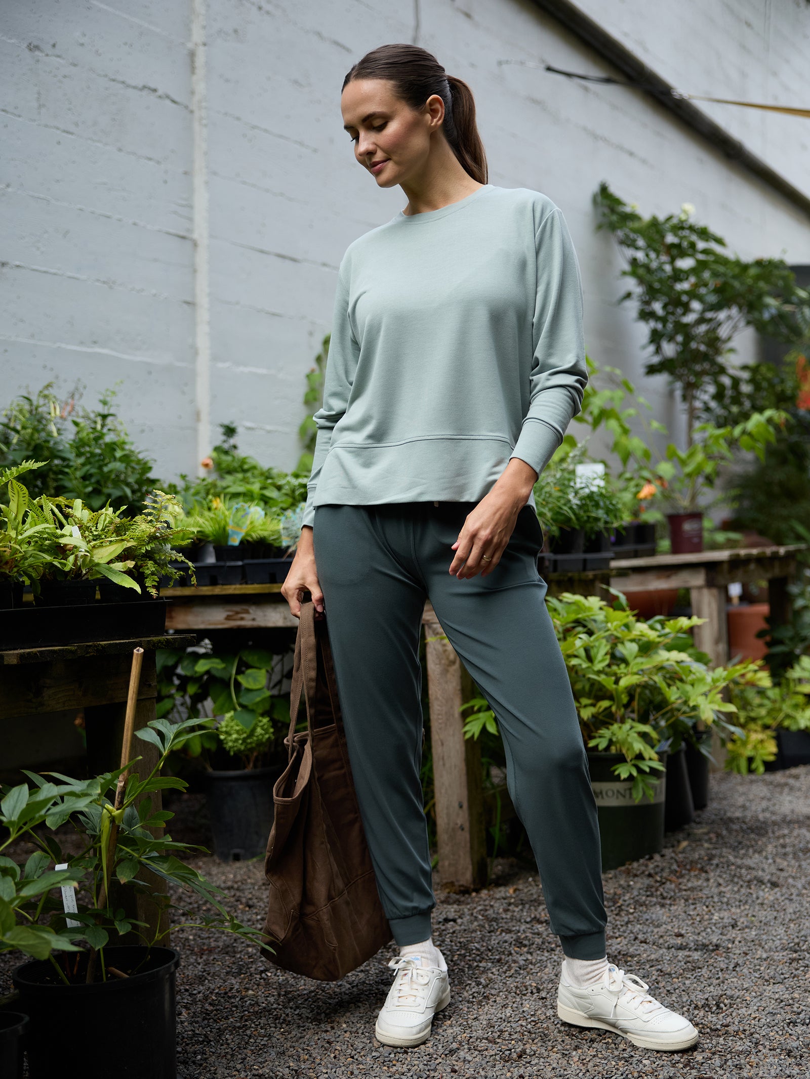 A woman stands outdoors in front of rows of potted plants, wearing a Cozy Earth Women's StudioLite Crewneck and matching track pants. She holds a brown tote bag in one hand and looks off to the side while smiling. She is dressed casually and wears white sneakers. 