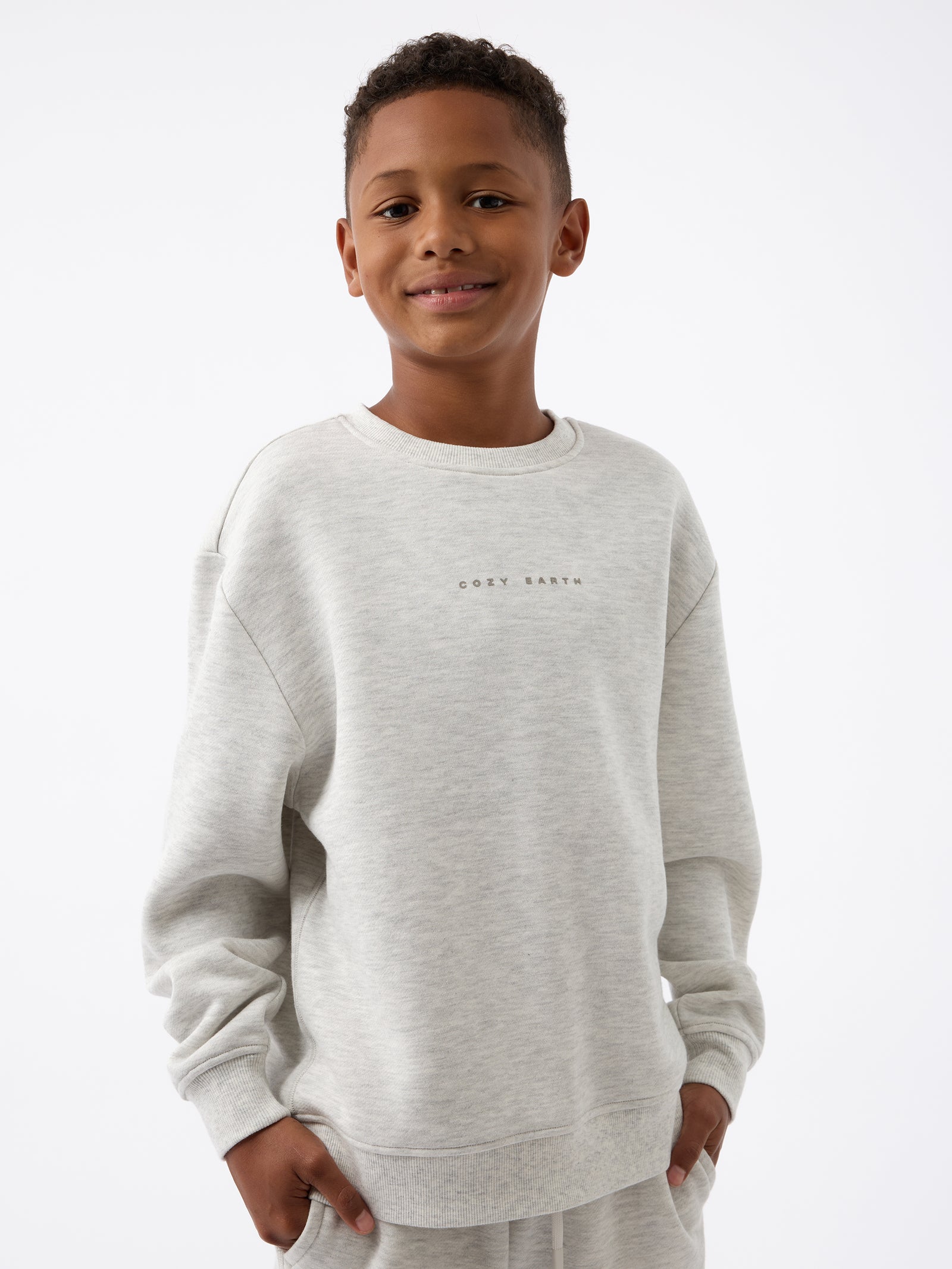 A smiling boy wearing the Kid's CityScape Crewneck by Cozy Earth, featuring "COZY EARTH" printed on it, stands against a plain white background. 