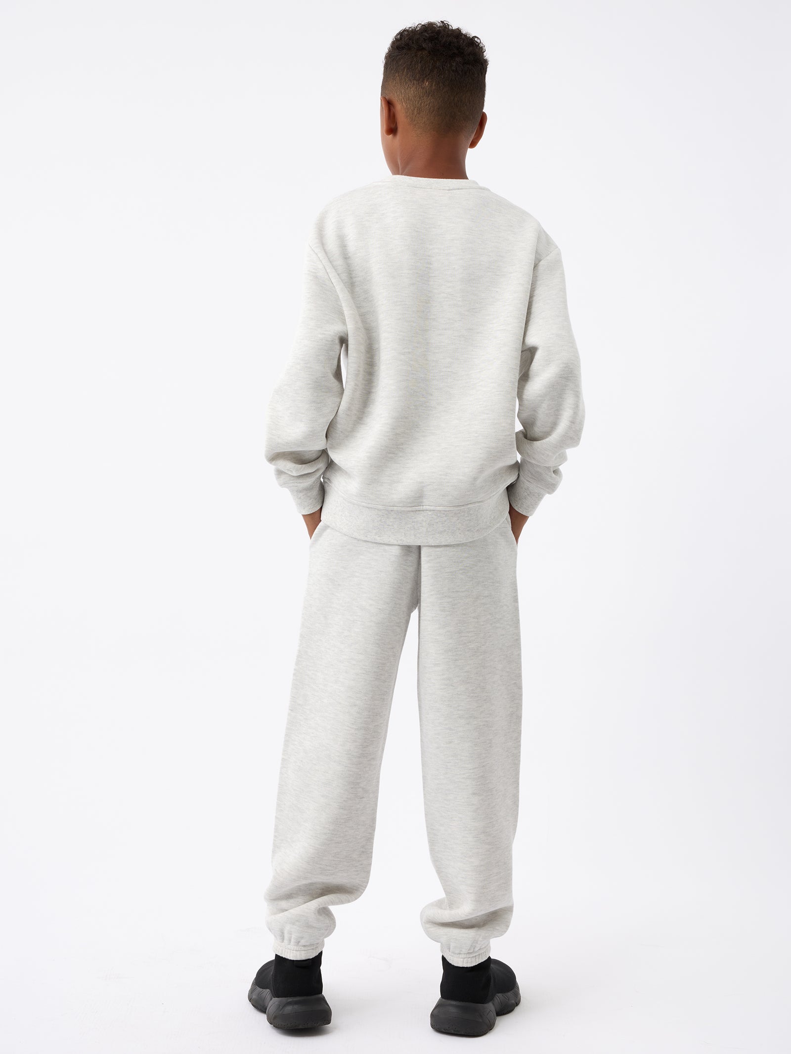 A person is standing against a plain white background, dressed in Cozy Earth's light gray Kid's CityScape Sweatpant with matching long sleeves. They face away from the camera, showcasing their short curly hair and black shoes. 