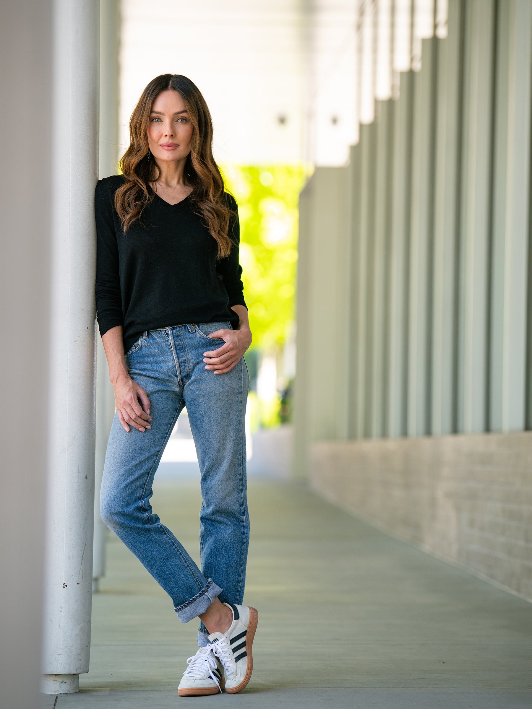A woman with long brown hair is standing outdoors in a modern architectural setting, leaning against a white column. She is wearing a black Cozy Earth Women's AirKnit V-Neck Sweater, blue jeans, and white sneakers with black stripes. The background includes vertical white panels and soft natural light. 