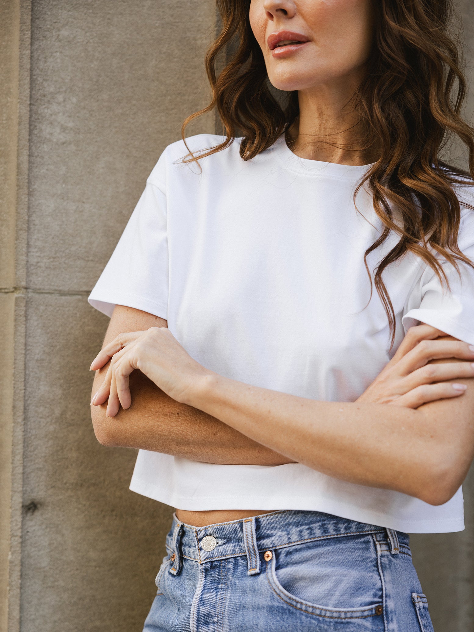 White All Day Cropped Tee. The photo of the All Day Cropped Tee is taken with a with a city style background and is worn by a woman. 