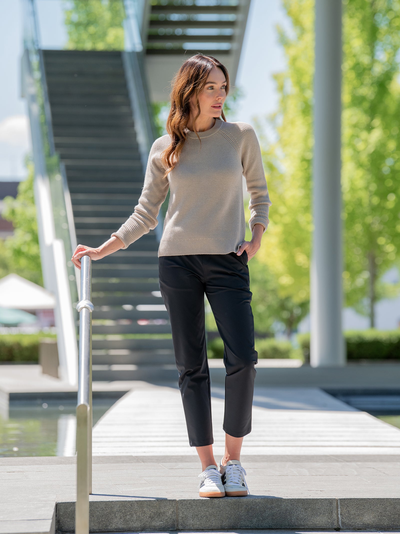 A woman stands outdoors on a staircase landing, holding onto a railing. She wears a beige sweater, Cozy Earth's Women's Always Cropped Pant in black, and white sneakers. Green trees and a modern staircase are in the background. She gazes away thoughtfully. 