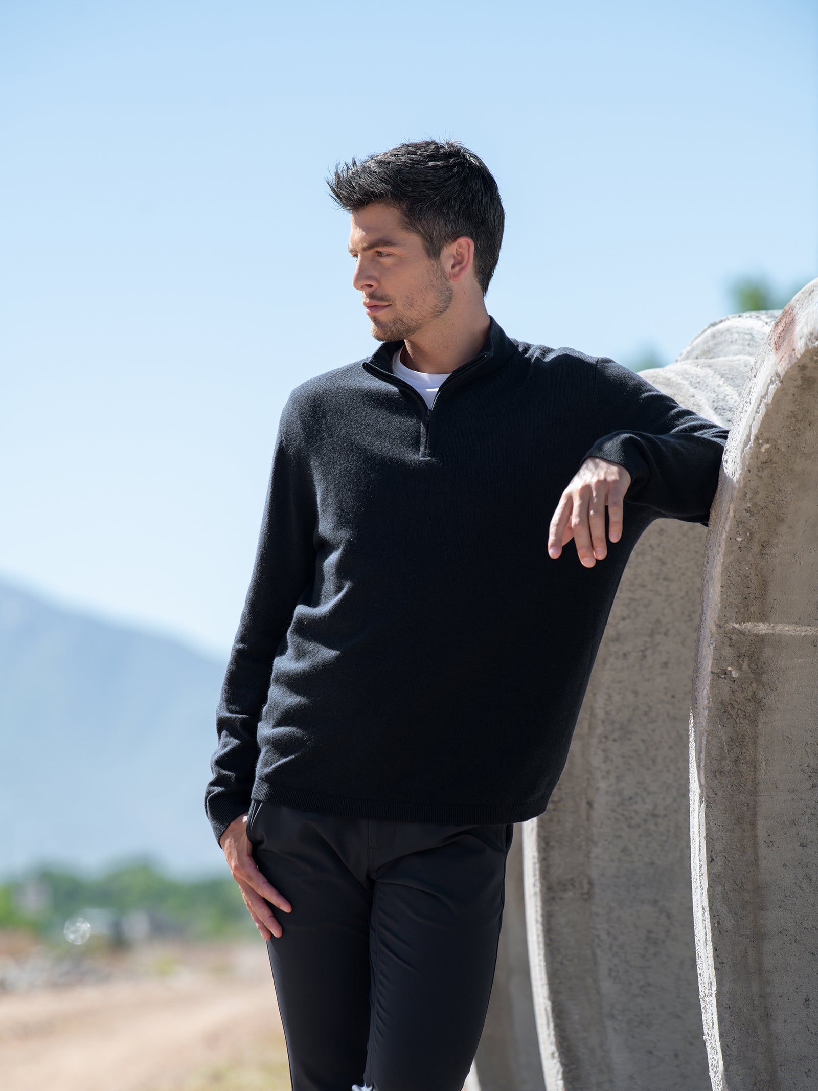 A man in Cozy Earth's Men's Quarter Zip Sweater and black pants stands outside during the day. He is leaning one arm on a large concrete pipe and looking down and to the side. There are mountains in the background and a bright blue sky above. 