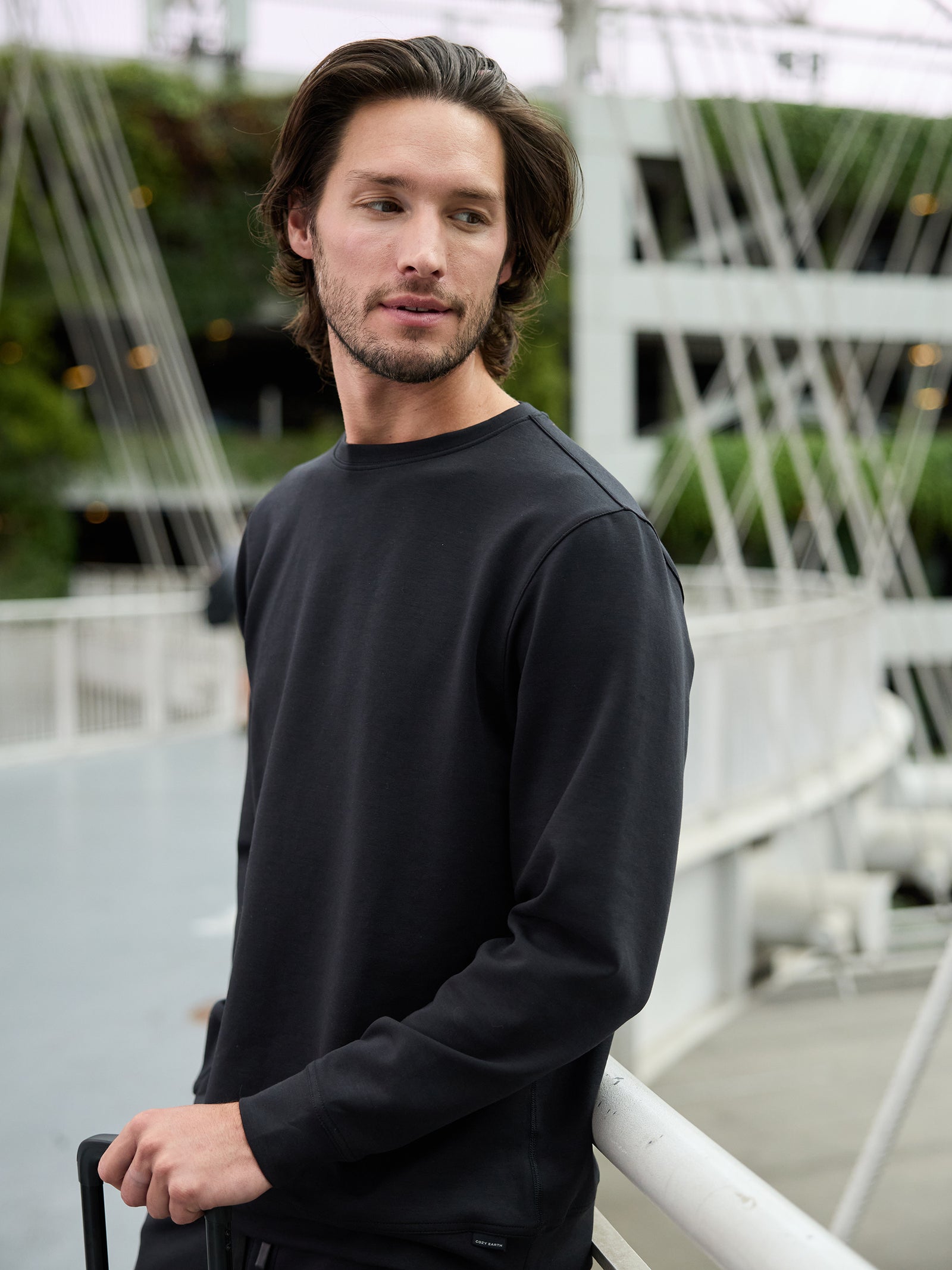 A man with medium-length brown hair and a beard is standing on a bridge, looking to his left. He is wearing the Cozy Earth Men's StretchTech Crewneck, a long-sleeved, dark-colored shirt, and has his left hand resting on a rail. The background includes greenery and modern architectural elements. 