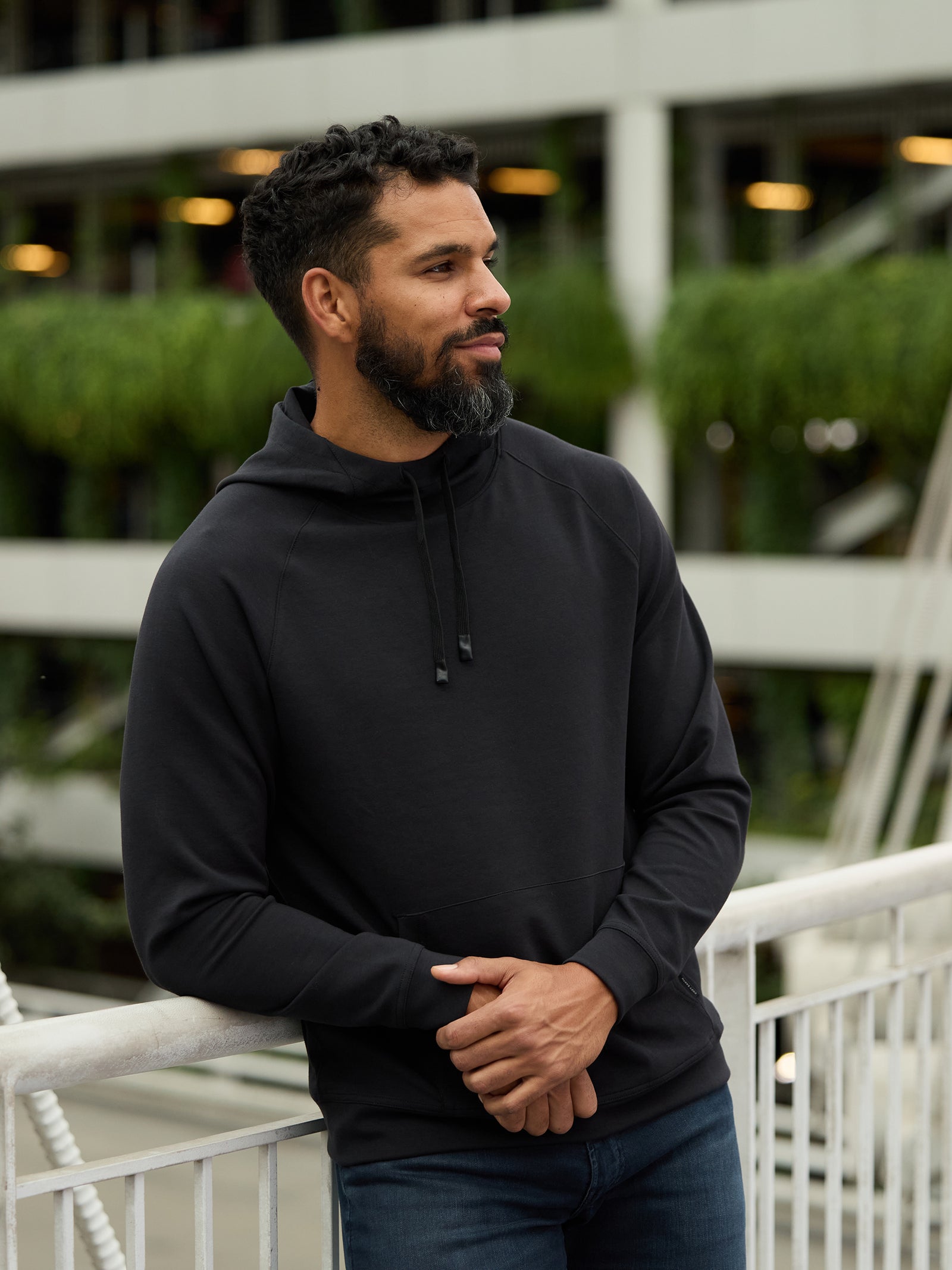 A bearded man with short curly hair stands outdoors, leaning on a white railing. He is wearing a black Men's StretchTech Hoodie by Cozy Earth and jeans, looking to the side thoughtfully. In the background, there is a building with greenery and multiple lit windows. 