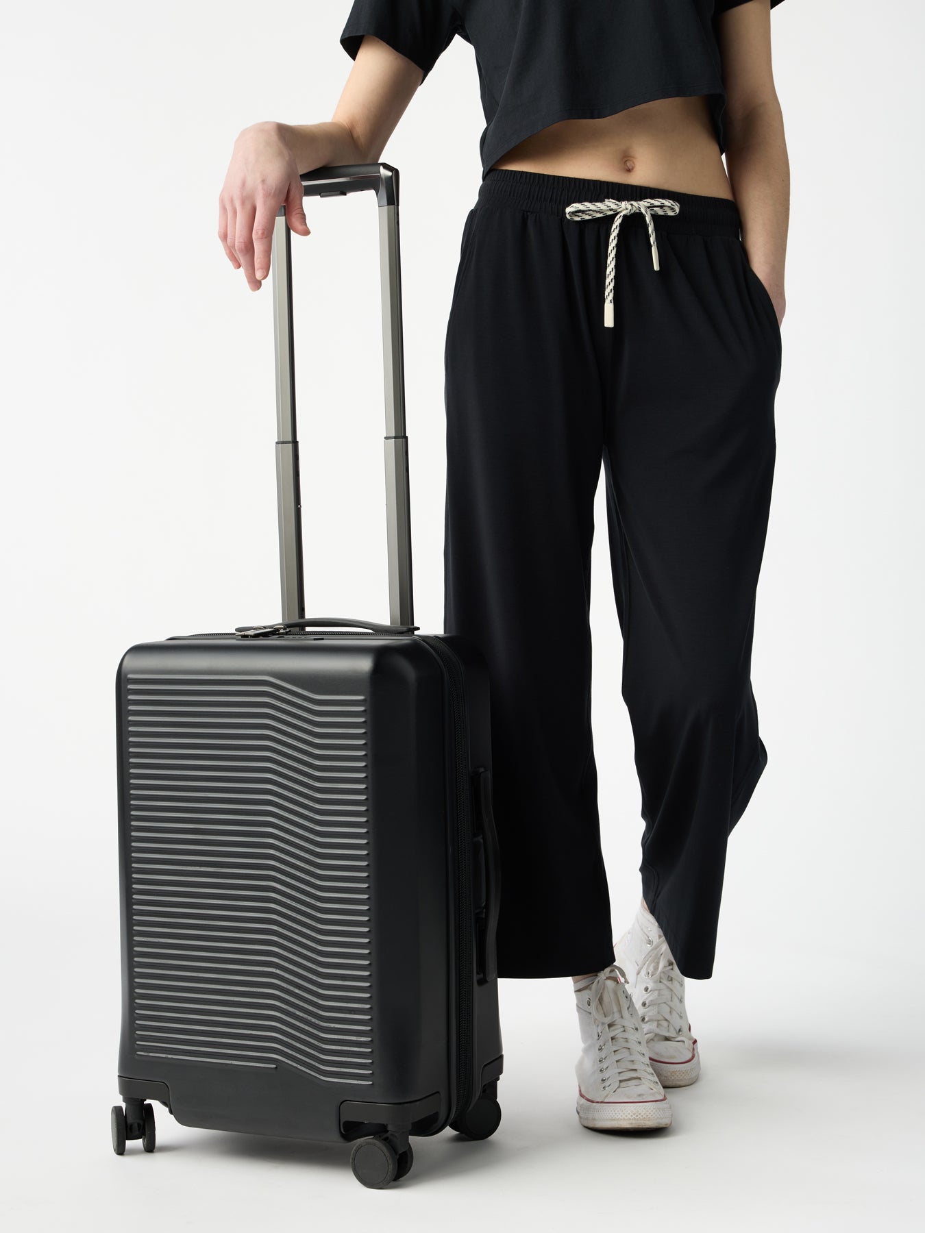 A person in a black crop top and Cozy Earth's Women's Studio Cropped Wide Leg Pant with white drawstrings stands holding the handle of a textured upright suitcase. They are wearing white sneakers against a plain white background. |Color:Jet Black