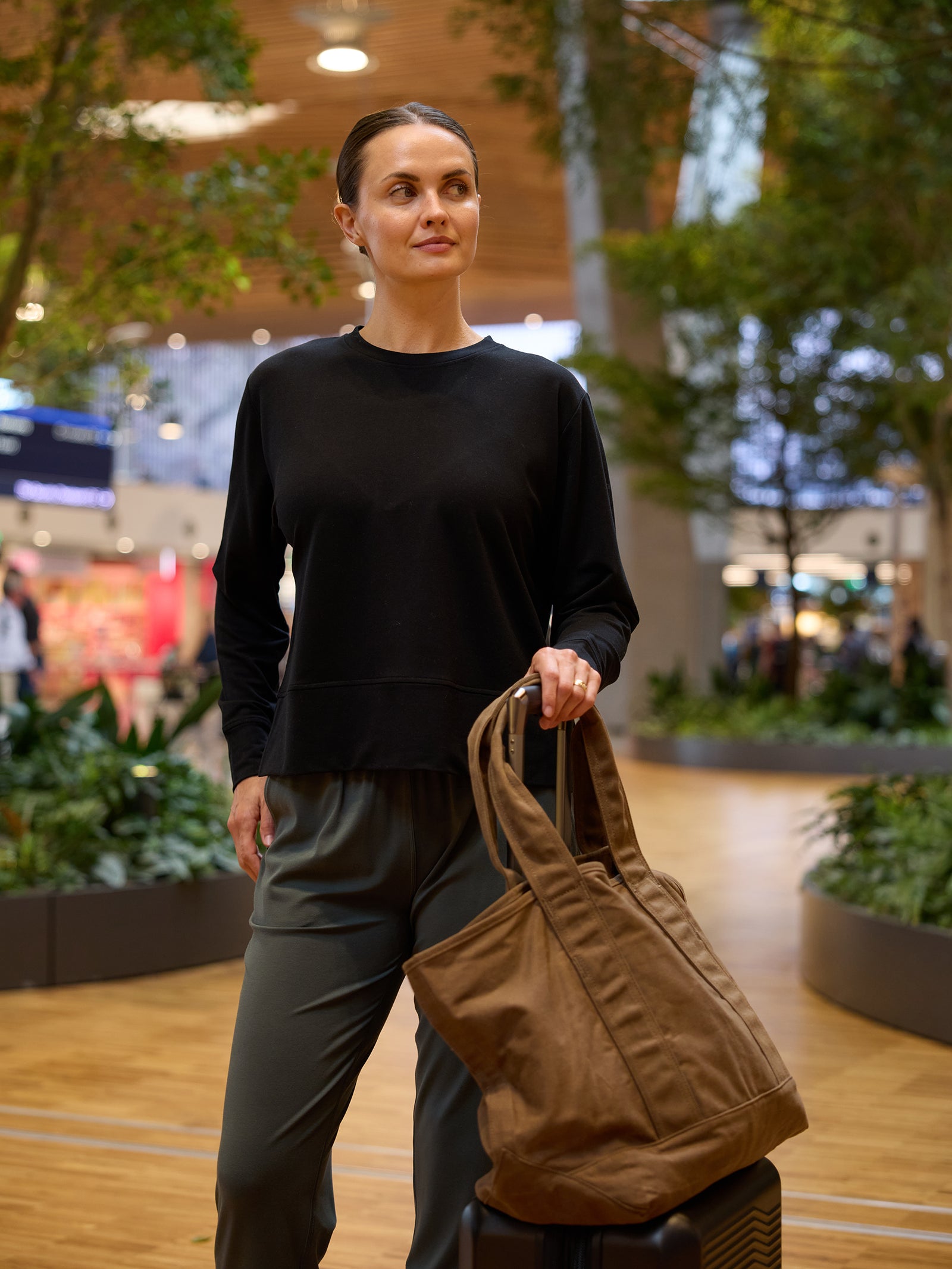 A woman stands in an indoor public space holding a large brown bag with one hand, which is resting on a suitcase. She is dressed in casual attire: Cozy Earth's Women's StudioLite Crewneck and gray pants. The background features greenery and out-of-focus people and shops. 