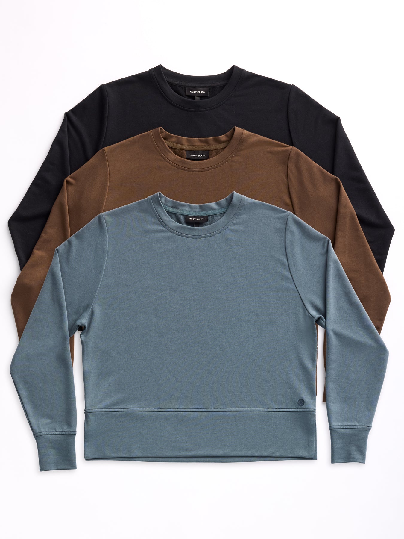 Three Women's StudioLite Crewnecks by Cozy Earth are stacked on a white background. The top one is black, the middle is brown, and the bottom is blue. Each features a simple round neckline and long sleeves.