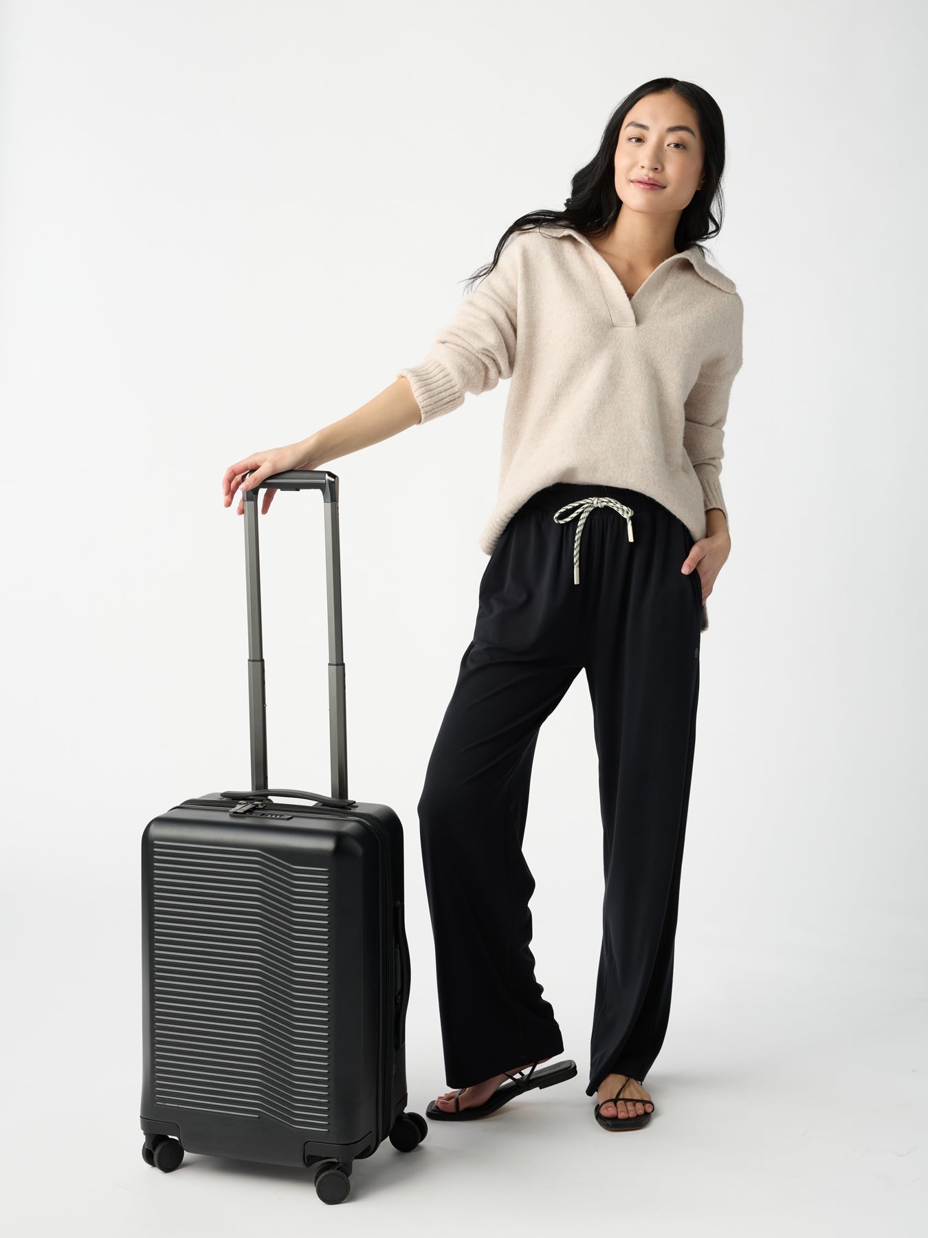 A person confidently stands against a plain white background in Cozy Earth's Women's Studio Wide Leg Pant, paired with a light beige sweater. One hand is in their pocket while the other holds a black suitcase handle. They're also wearing black sandals. 