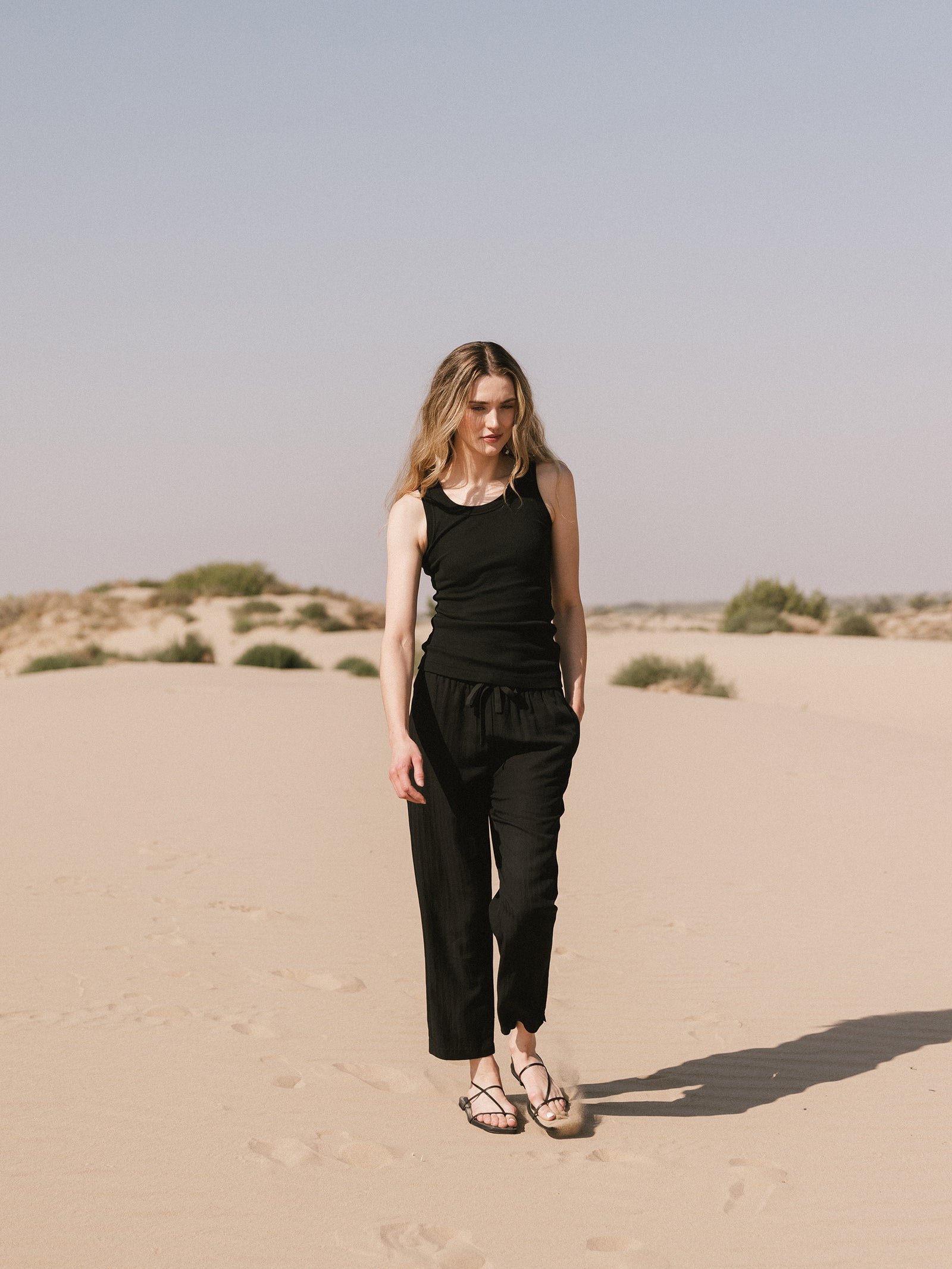 A woman with long blonde hair walks barefoot on a sandy desert wearing a black tank top and Cozy Earth's Women's Sunset Cropped Pant. She looks down, and her shadow stretches beside her under the clear sky. Sparse desert vegetation is visible in the background.