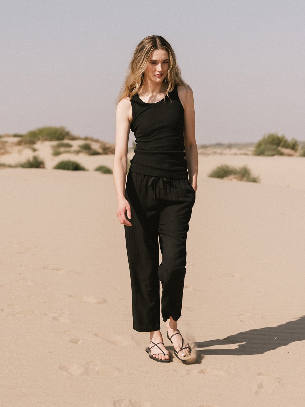 A woman with long blonde hair, wearing Cozy Earth's Women's Sunset Cropped Pant and a black sleeveless top, strolls on sandy terrain with sparse shrubs under a clear sky. 