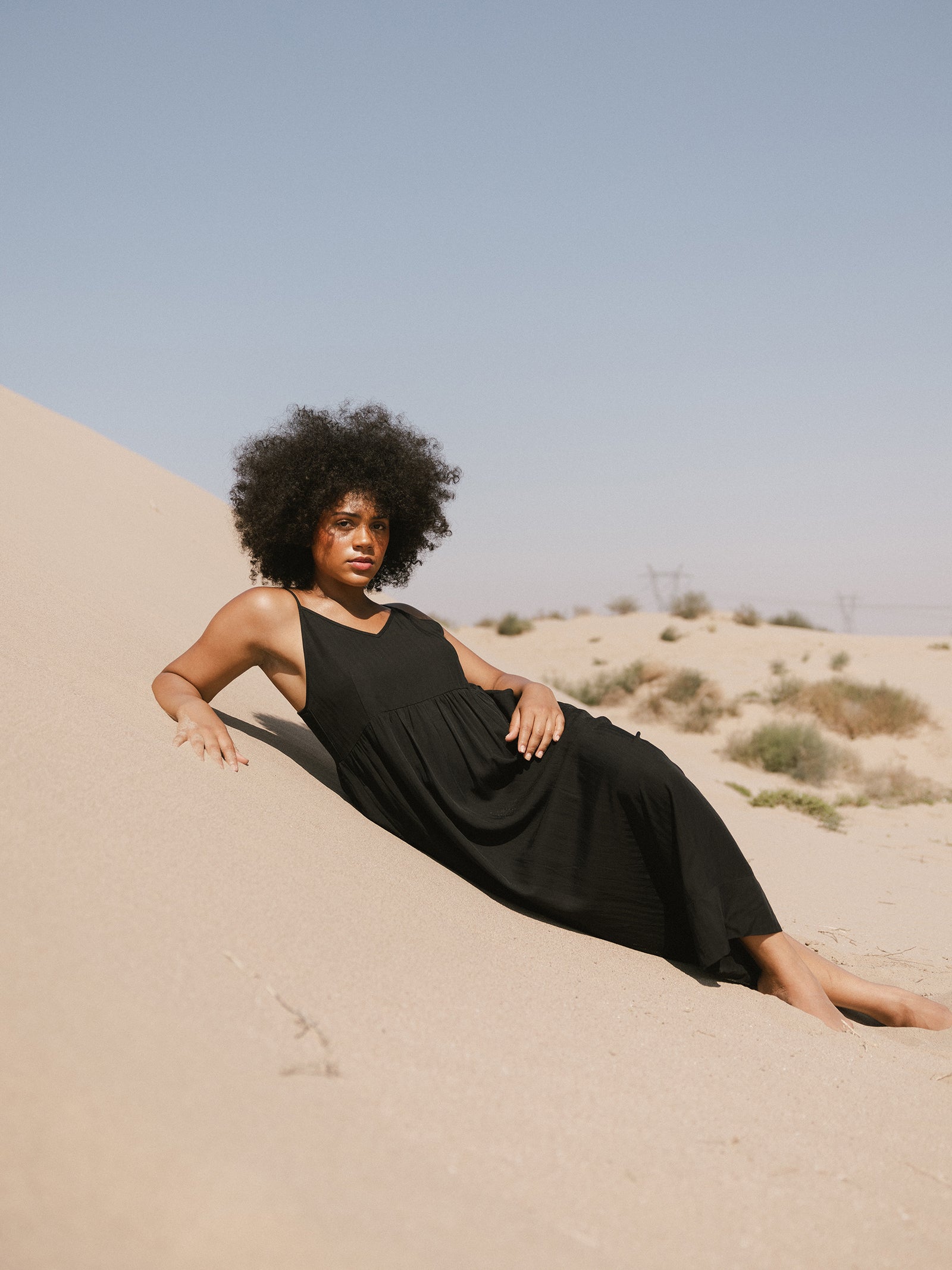 A woman with an afro hairstyle is reclining on a sandy dune, wearing a Cozy Earth Women's Sunset Gathered Dress. She is looking at the camera with a calm expression. The background features a clear sky and sparse vegetation on the sand. 