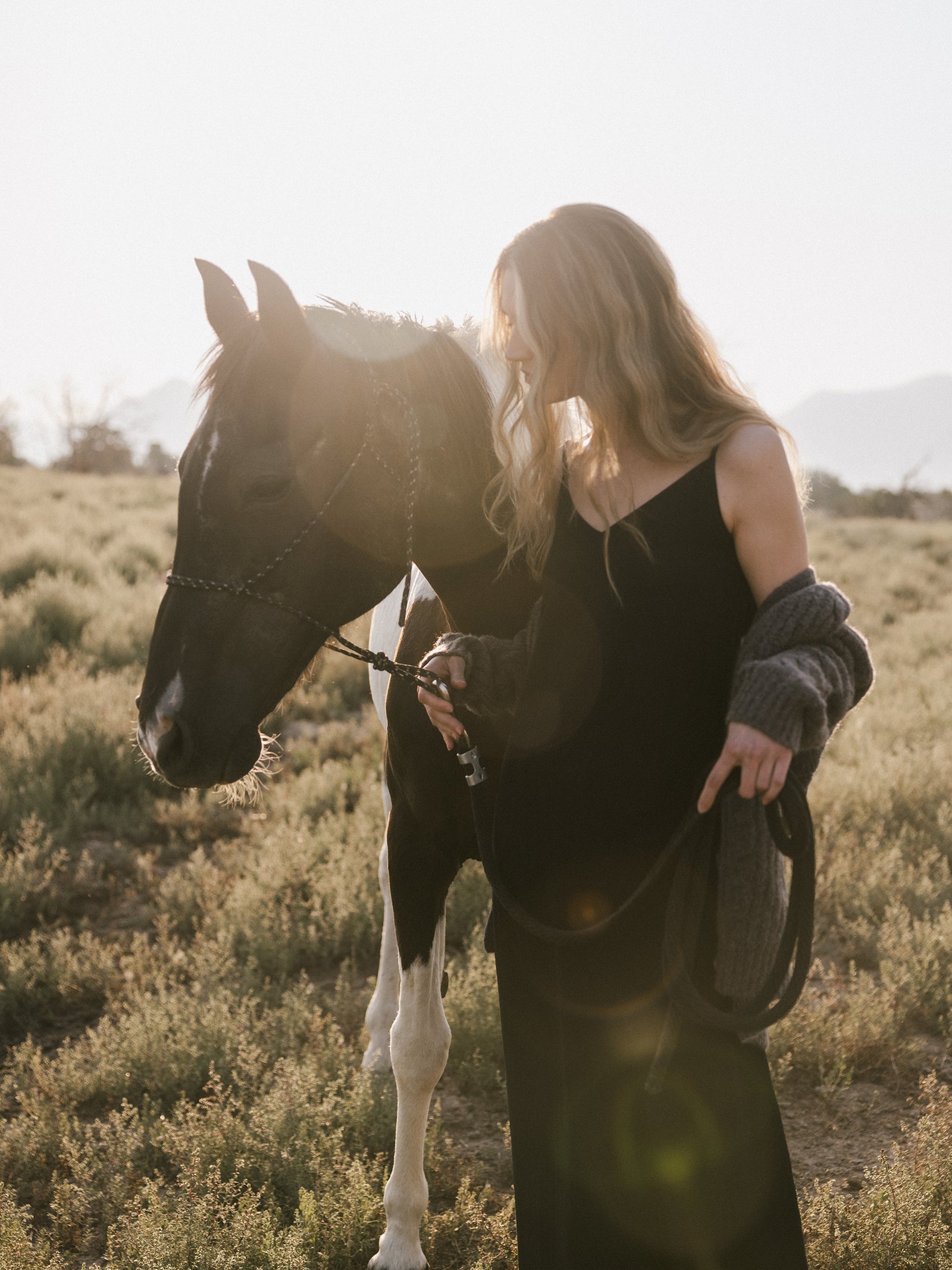 A woman wearing the Women's Sunset Slip Dress by Cozy Earth and a gray sweater stands in a grassy field at sunset, gently holding the reins of a black and white horse. With their heads slightly bowed, sunlight filters through the scene, casting a warm glow. 