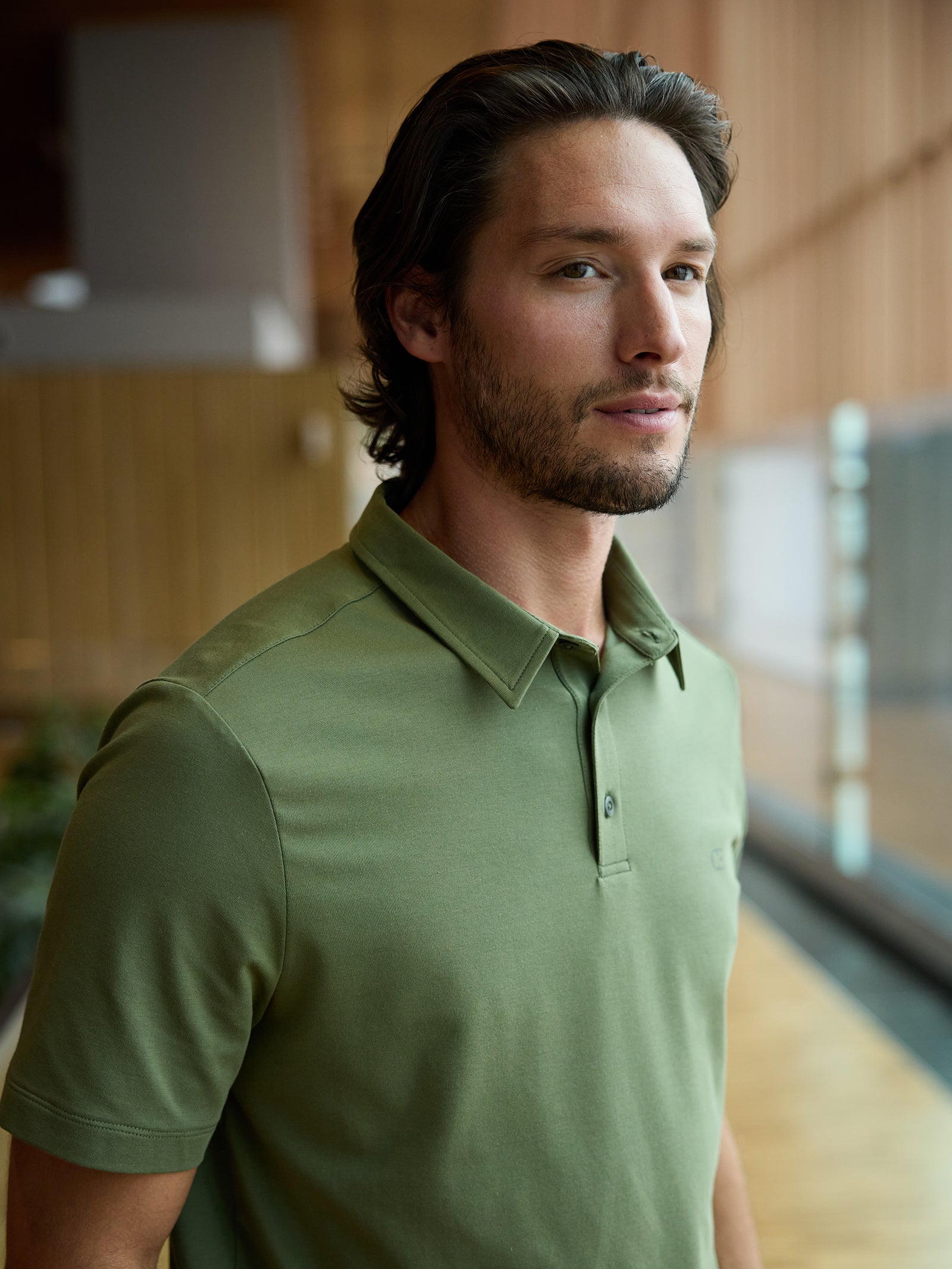 A man with shoulder-length dark hair and a beard is wearing a green Men's Everyday Polo from Cozy Earth. He stands indoors against a wooden wall, looking slightly to the side in a warm and softly lit setting. 