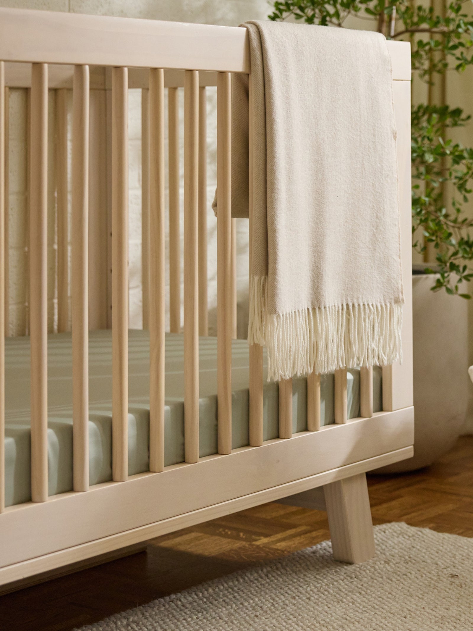 The Cozy Earth Crib Sheet by Cozy Earth is draped over a wooden crib with vertical slats, placed on a hardwood floor with a beige rug. A green plant adds a natural touch to the serene room. 