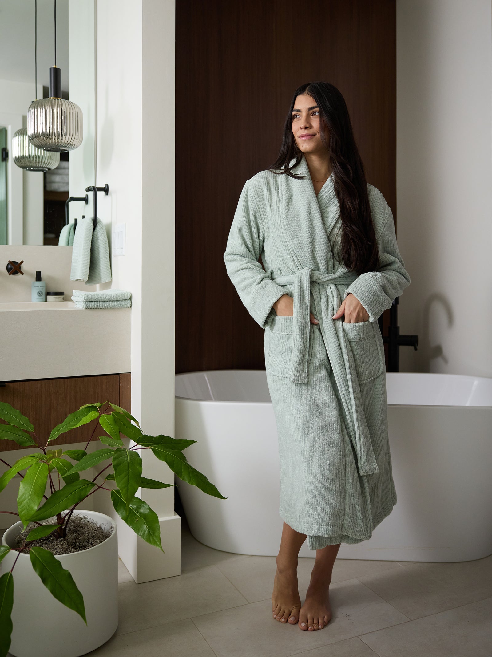 A person with long dark hair stands barefoot in a bathroom, wearing the Cozy Earth Ribbed Terry Bath Robe in light green. The person is in front of a bathtub and beside a vanity with a potted plant and neatly arranged towels. A modern light fixture is visible on the bathroom wall. 