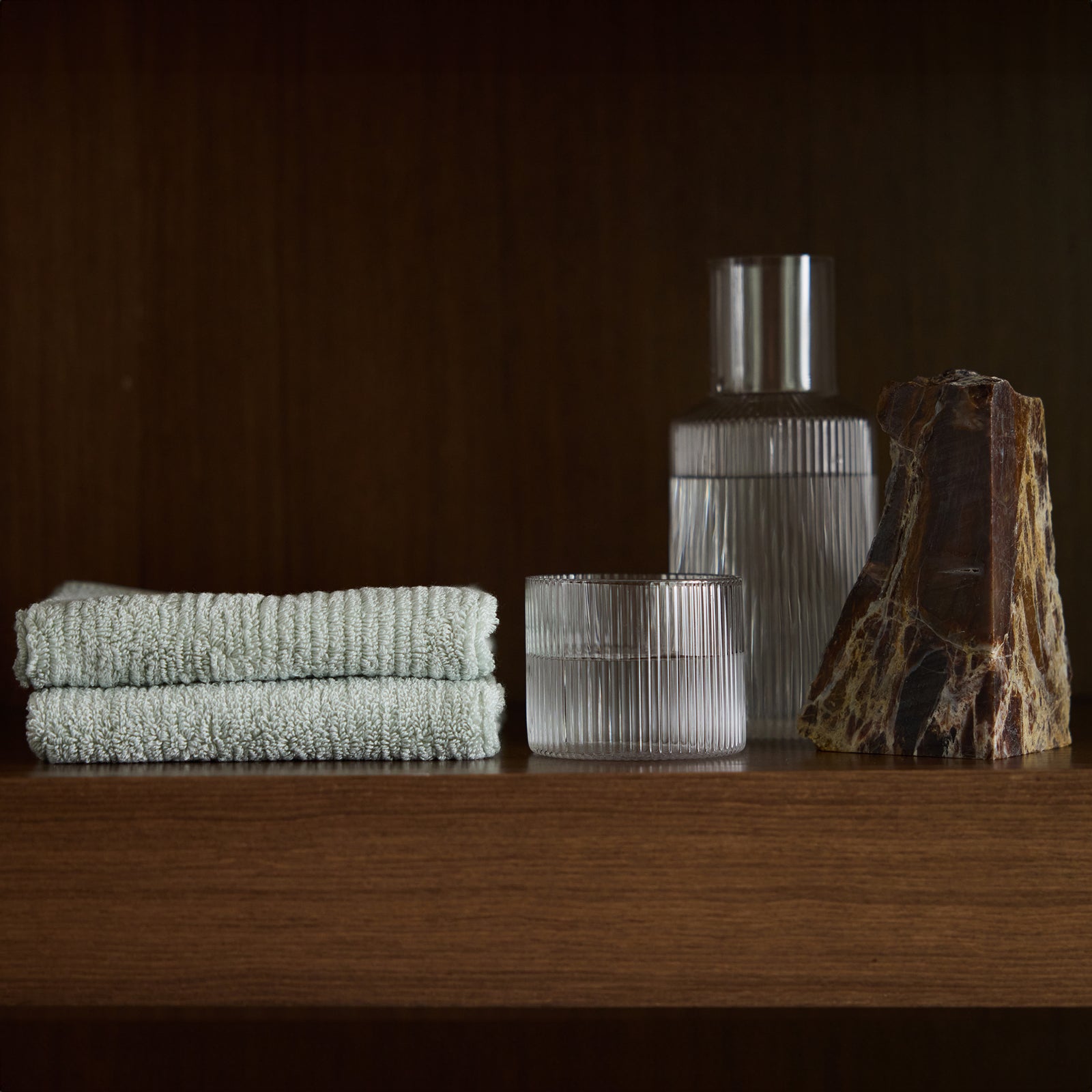 A wooden shelf displaying a neatly folded stack of light green Ribbed Terry Washcloths from Cozy Earth, two clear ribbed glass containers, and a decorative stone with brown and beige patterns. The dark brown background accentuates the items on the shelf. 