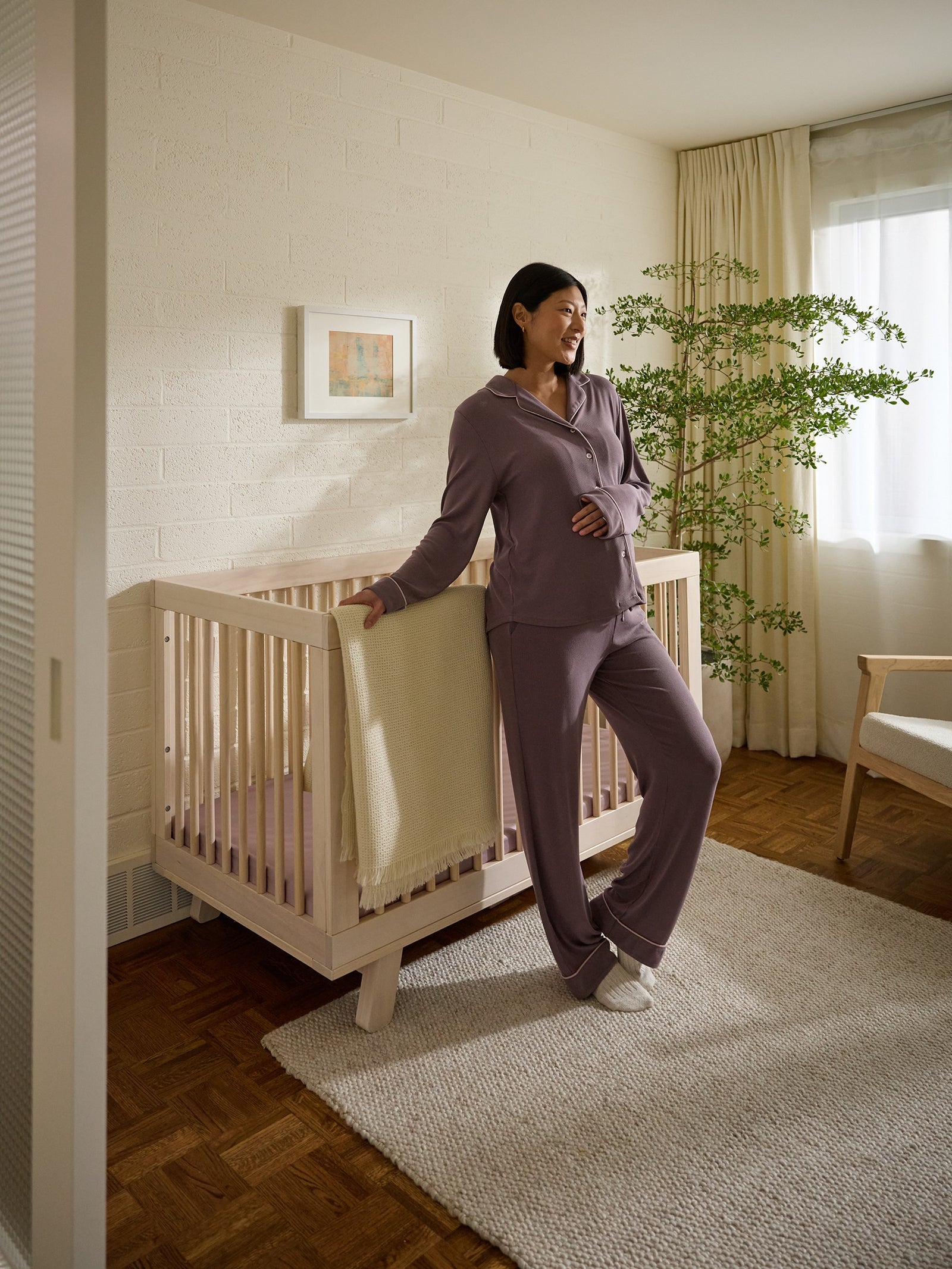 A woman in purple pajamas stands beside a wooden crib adorned with a Cozy Earth Crib Sheet in a cozy room. She smiles, resting her hand on her baby bump. The space features a leafy plant, framed picture, and light streaming through a curtained window. 