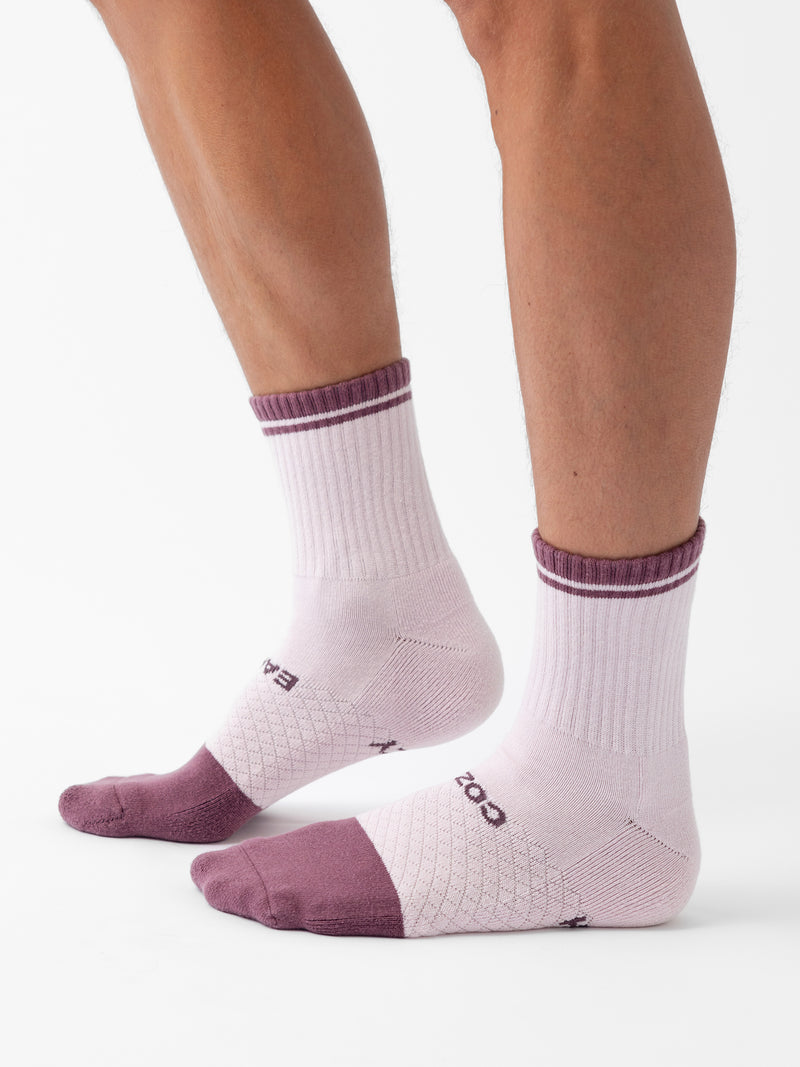 Two legs are shown wearing a pair of Cozy Earth's Essential Quarter Sock from the 2-Pack. The white socks feature a purple heel, toe, and top trim, with text and geometric patterns over the instep. The background is plain white.
