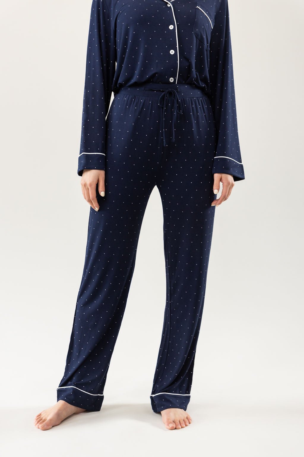 A person is shown barefoot in a Cozy Earth's Women's Bamboo Stretch-Knit Long Sleeve Pajama Set featuring a navy blue color with small white polka dots, white piping, a button-up top, and drawstring waist pants against a plain light background. 