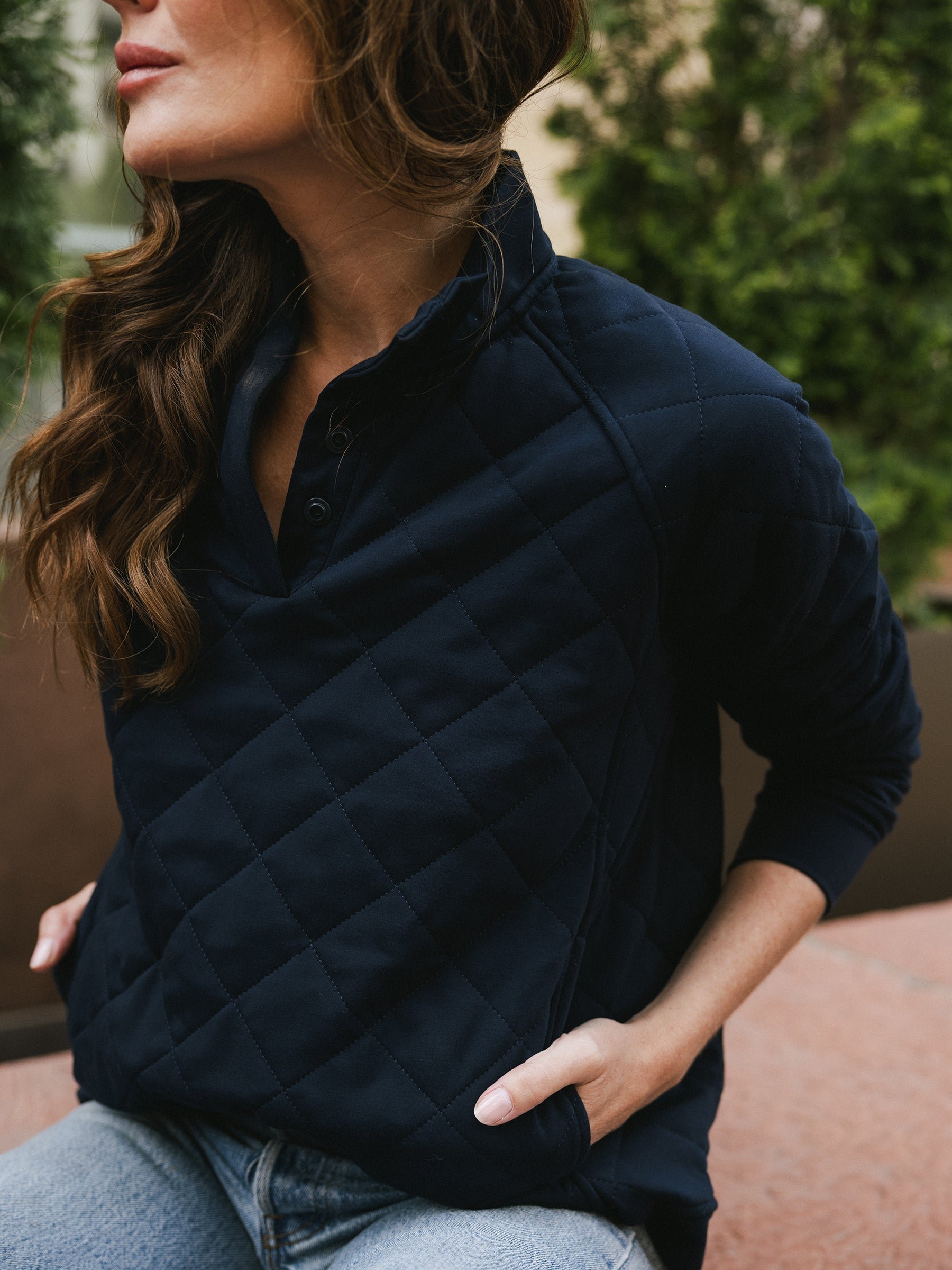 A person with wavy brown hair is wearing a Women's Ultra-Soft Bamboo Quilted Snap Pullover by Cozy Earth in dark blue and light blue jeans. They are sitting with one hand in their jeans pocket and the other resting on a bench, with greenery visible in the background. 