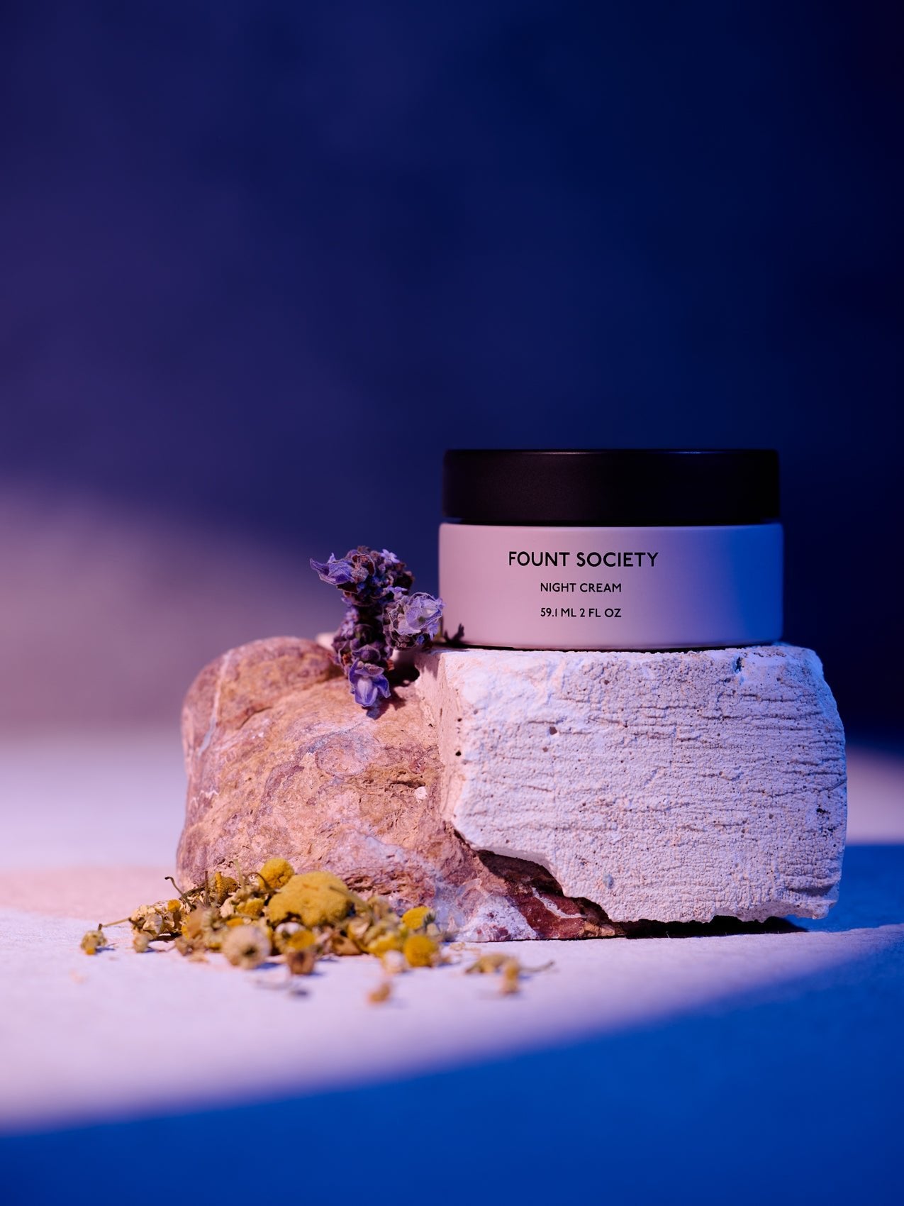 A jar of Cozy Earth Night Cream rests on a textured stone, surrounded by small dried flowers under gentle purple and pink lighting. |Size:2 OZ